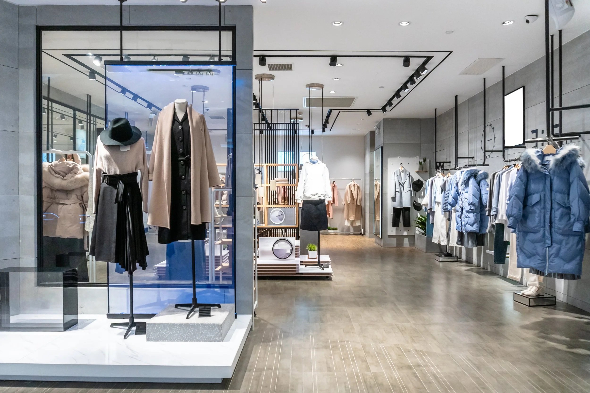 A clothing store with various winter jackets, coats, and accessories on display. Mannequins dressed in winter clothing stand in front of the store window. The interior has modern decor with track lighting and shelves with small potted plants.