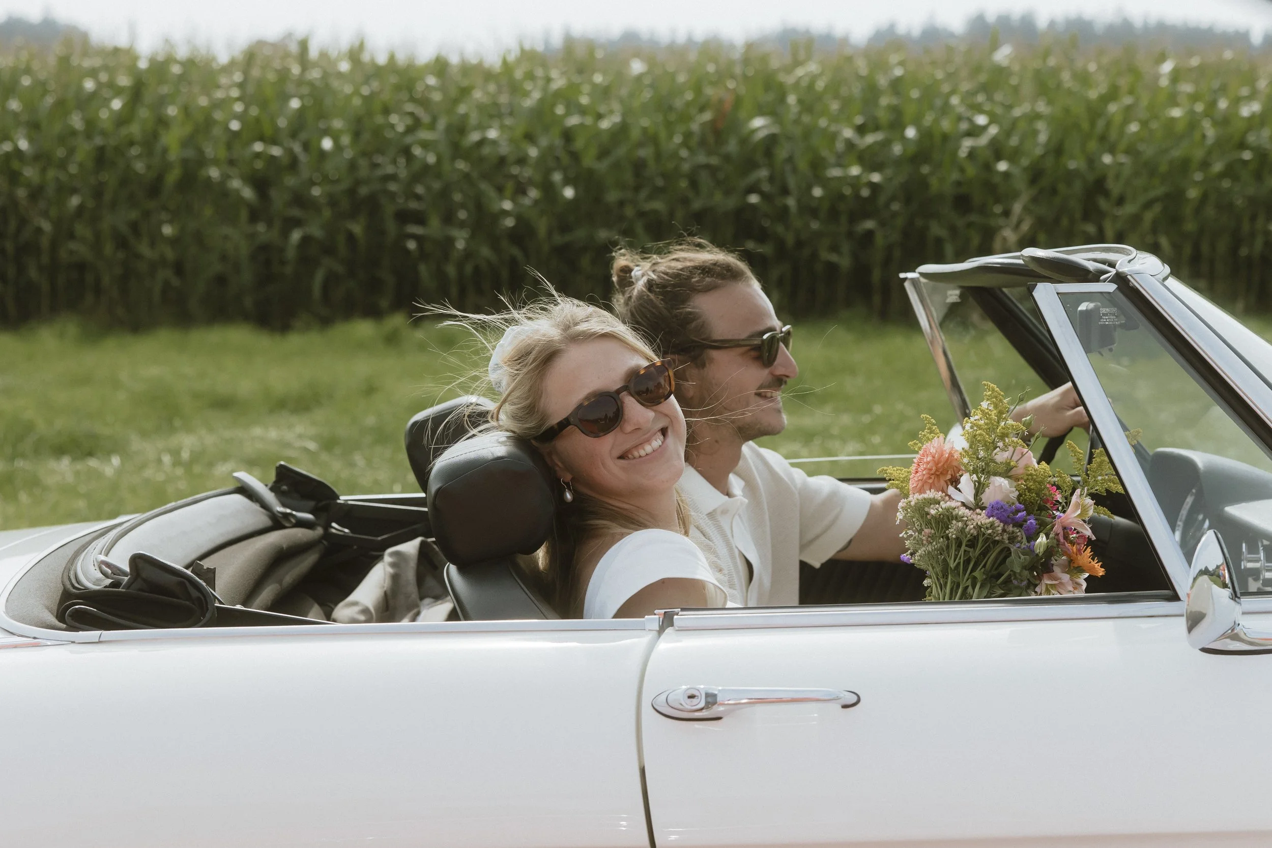 Ein lächelndes Paar in einem weißen Cabrio fährt durch eine ländliche Gegend, der Fahrer hält einen Blumenstrauß. Im Hintergrund ist ein Maisfeld zu sehen.