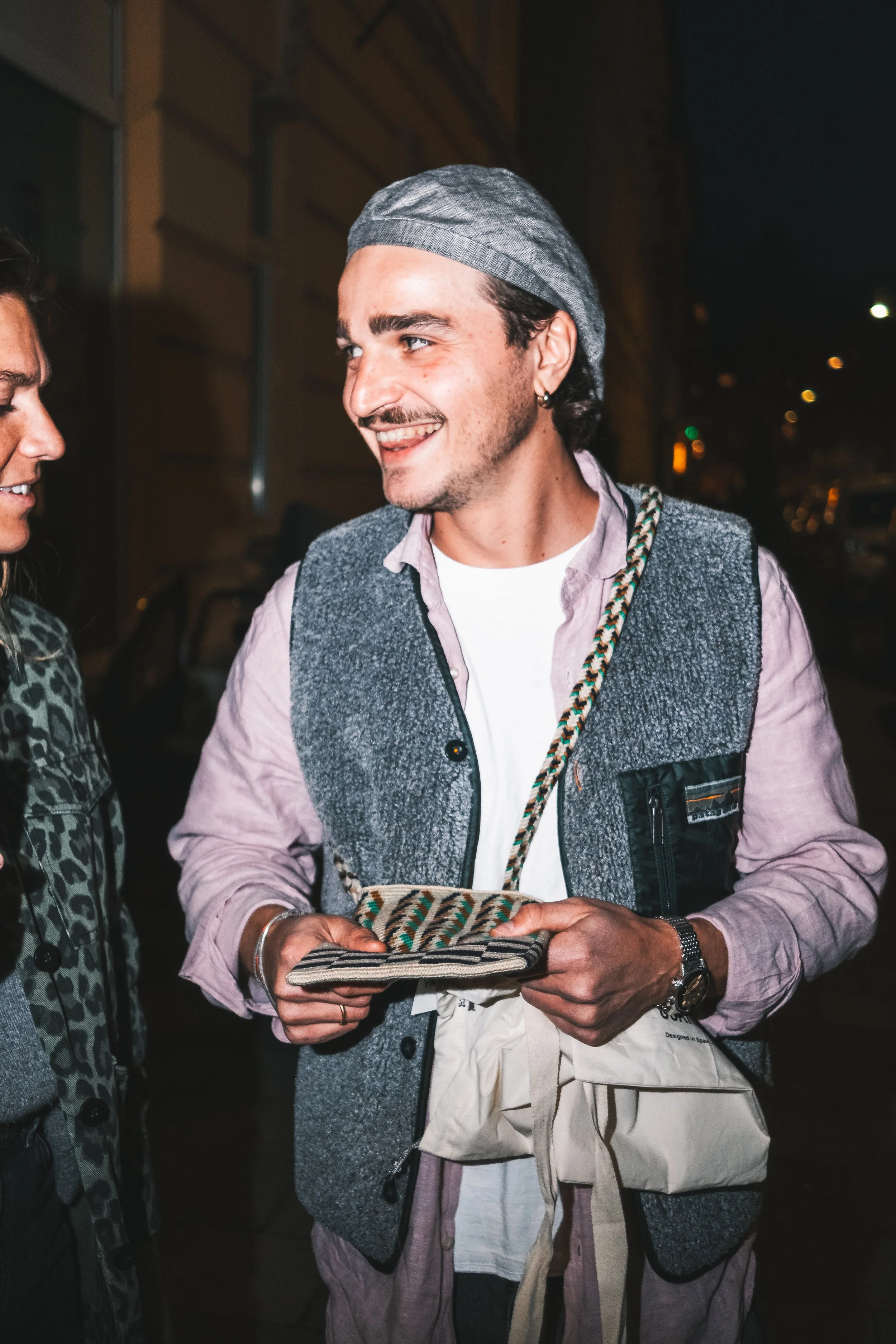 Ein lächelnder Mann in lässiger Kleidung hält eine Tasche in der Hand und unterhält sich mit einer anderen Person auf einer nächtlichen Straße.