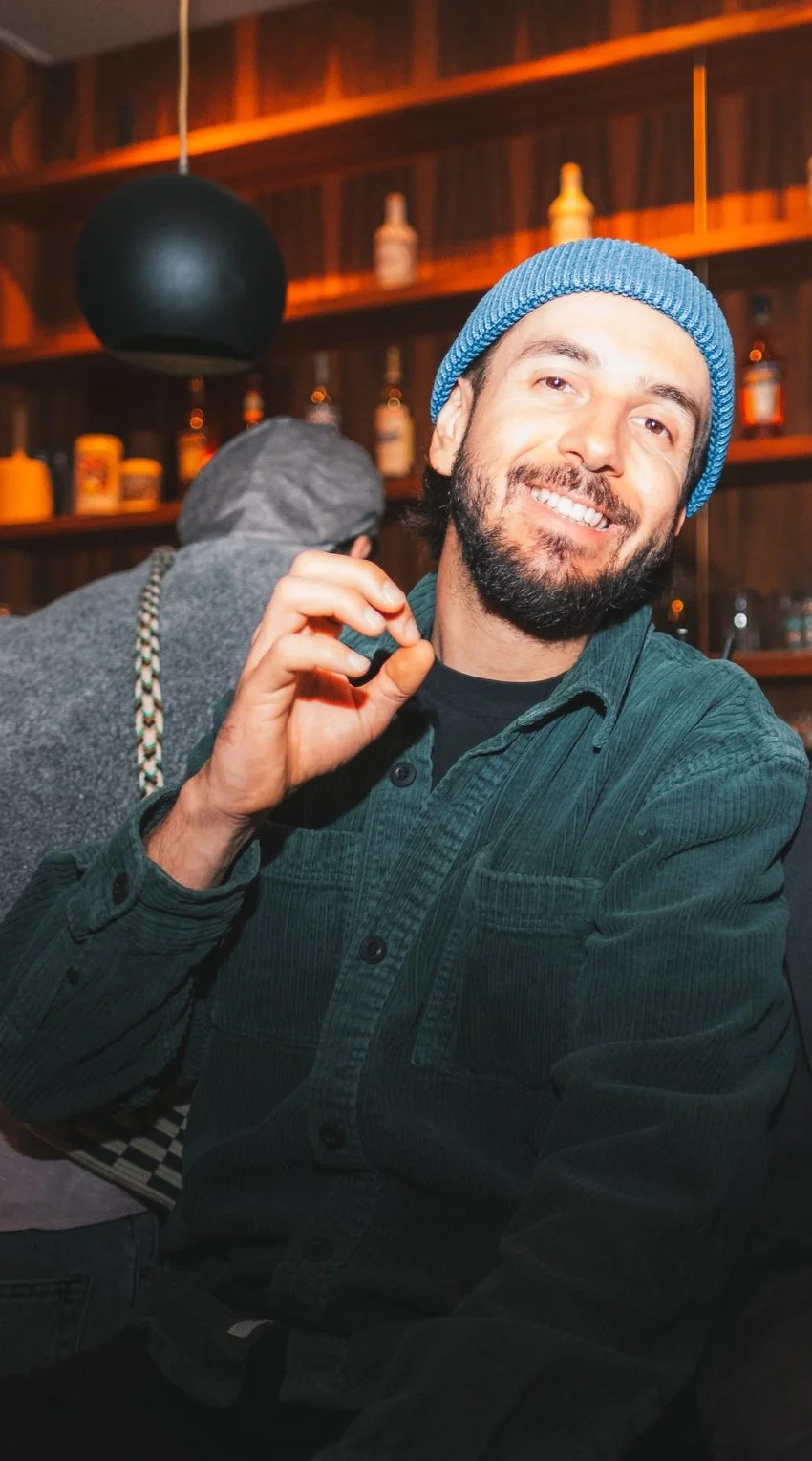Mann mit blauem Mütze und grünem Hemd lächelt in die Kamera, im Hintergrund Regale mit Flaschen.