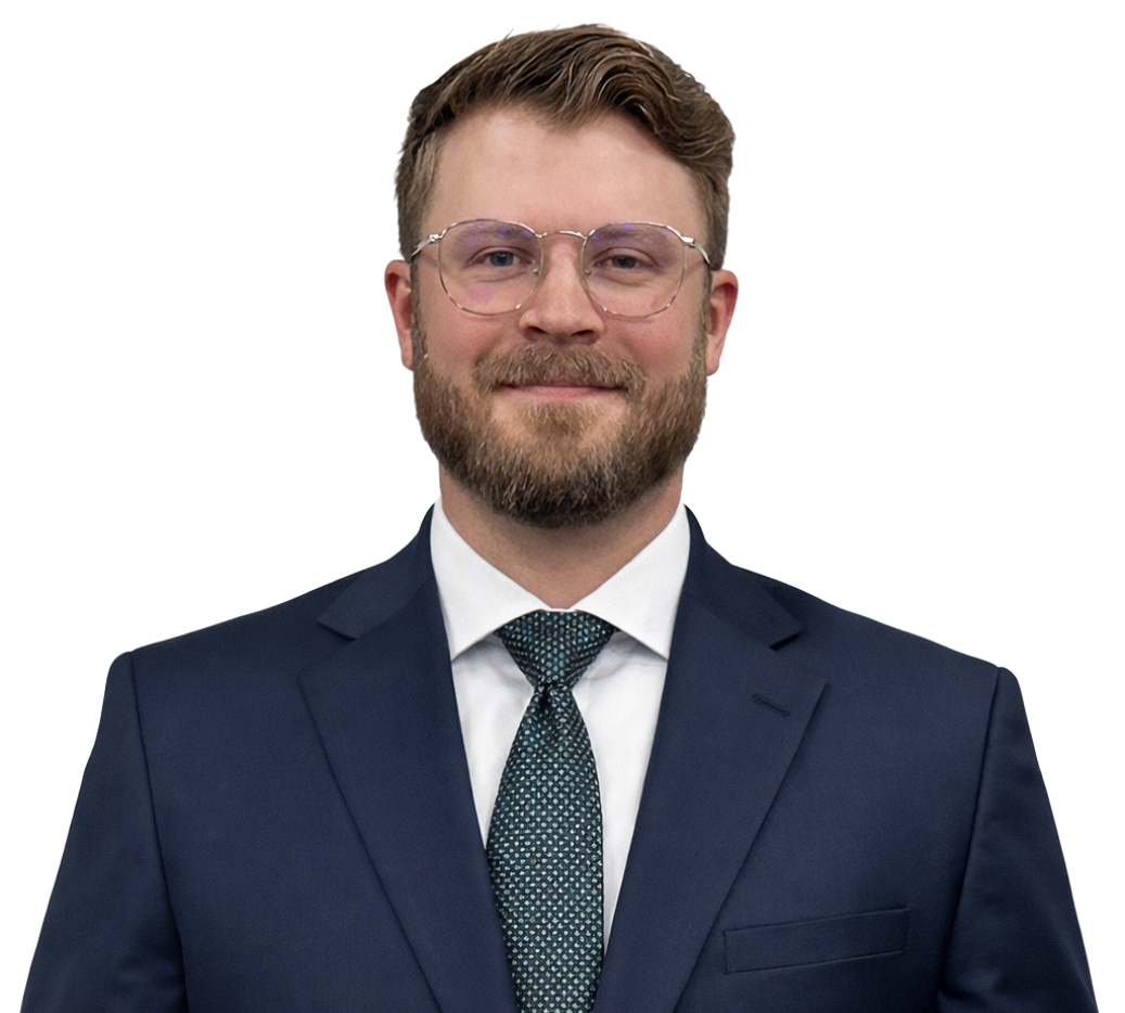 Portrait of a young man with glasses, a beard, and light brown hair, wearing a navy suit, white shirt, and patterned tie, smiling against a white background.