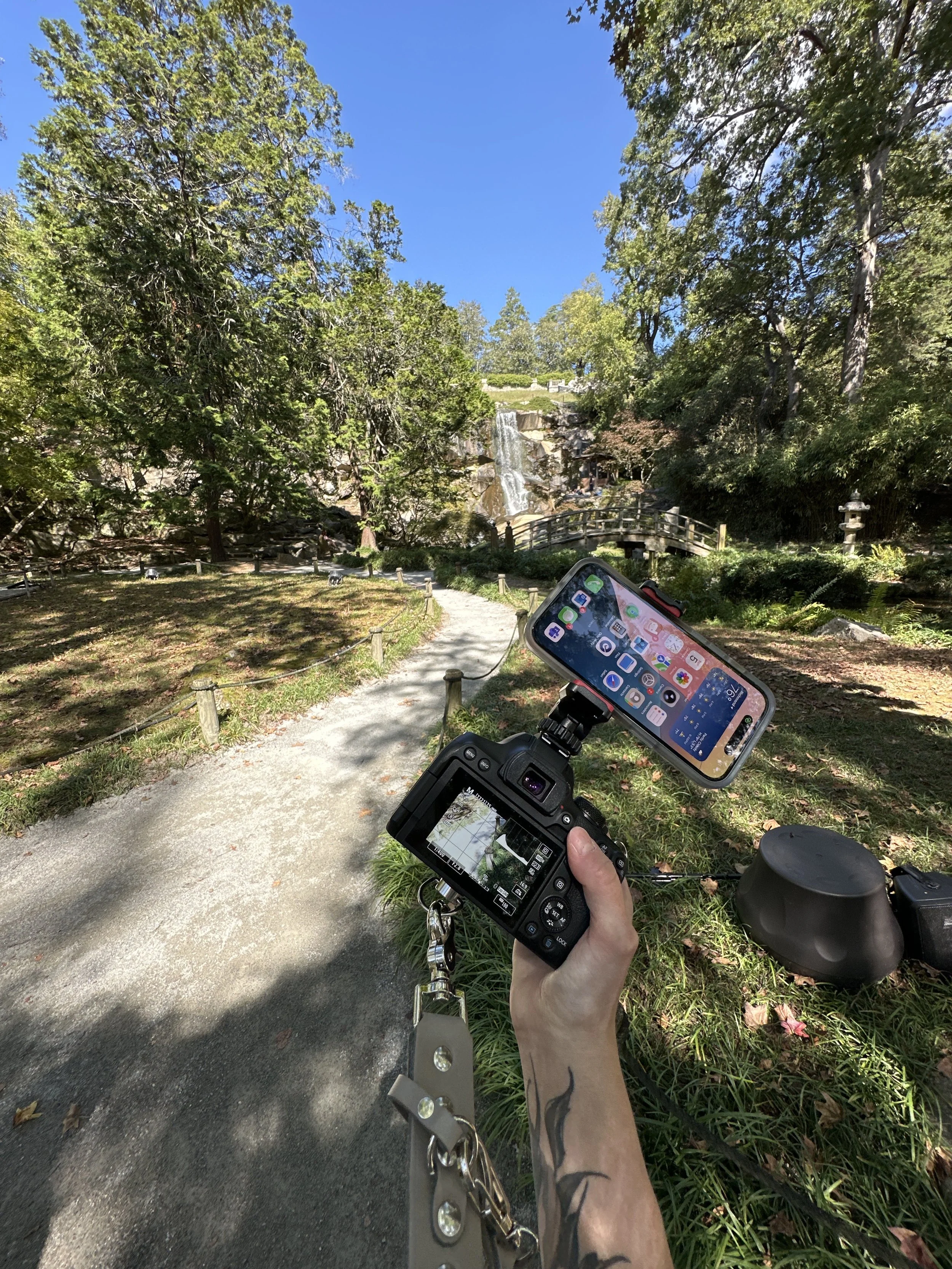 A person taking a photograph with a digital camera and smartphone in a park with a waterfall and lush greenery in the background.
