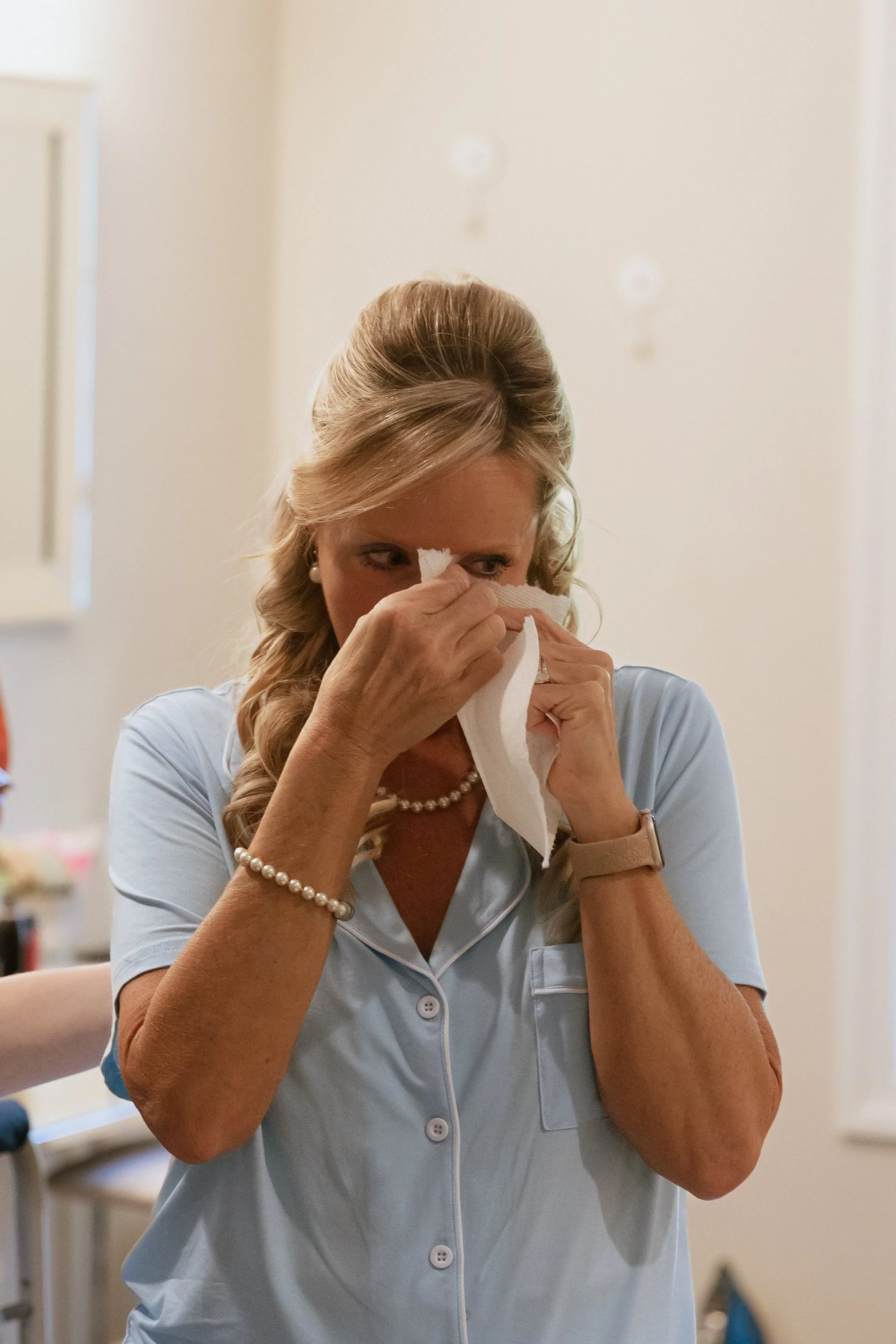 A woman with blonde hair, wearing a pearl necklace and bracelet, dressed in light blue pajamas, appears to be wiping her eyes with a tissue.