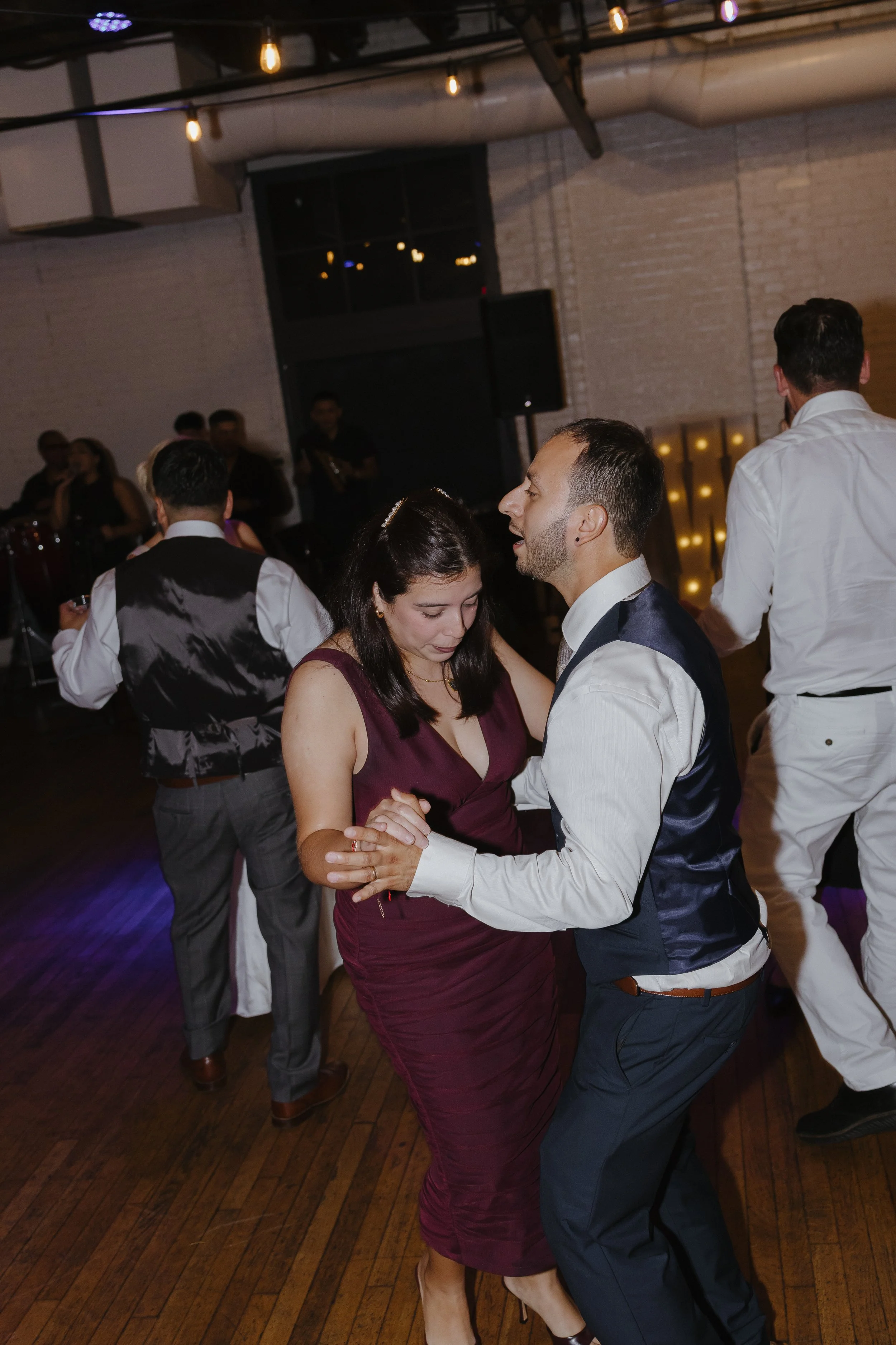 People dancing at a wedding reception in a dimly lit venue, with a woman in a burgundy dress and a man in a white shirt and dark vest dancing closely.