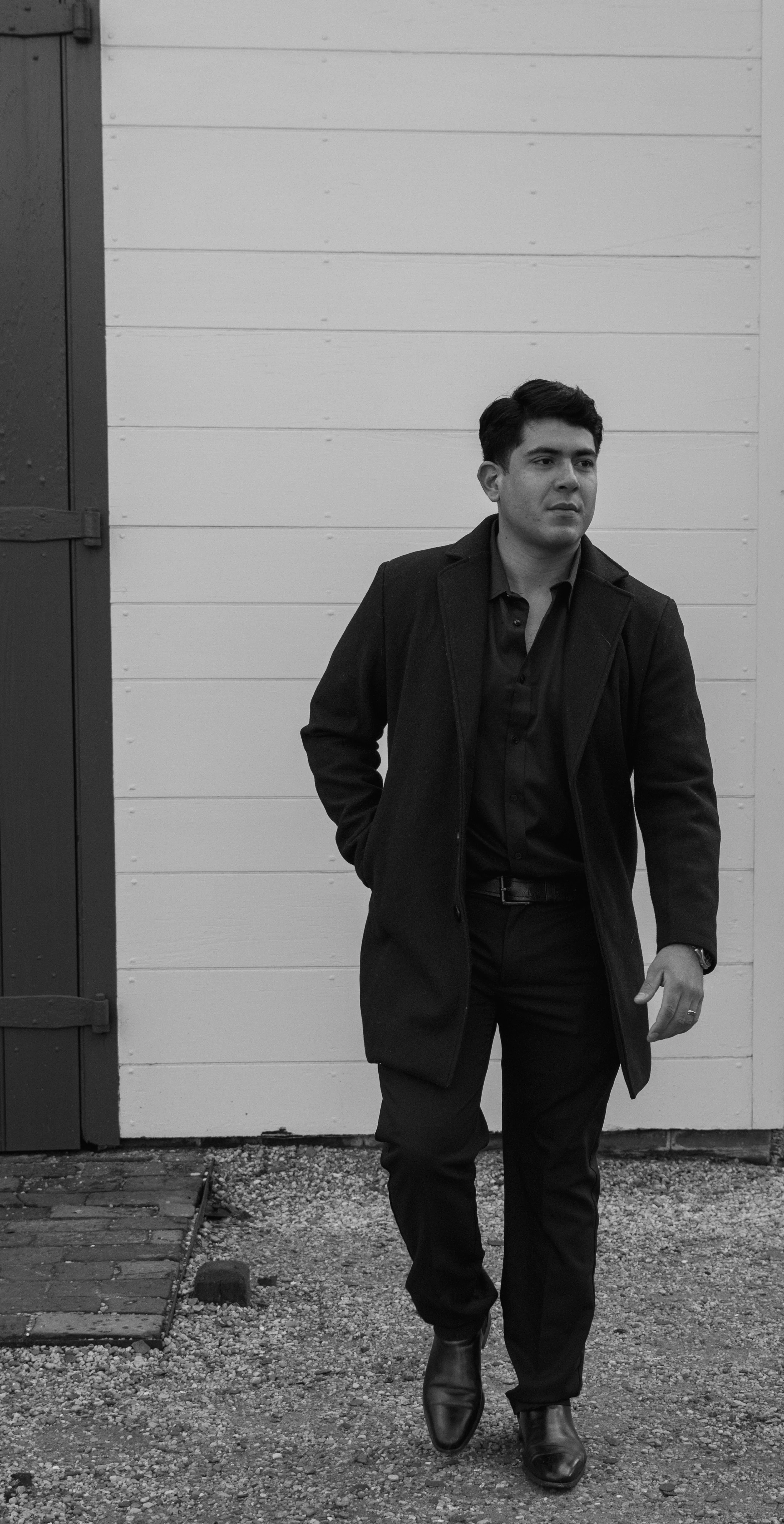 Black and white photo of a man in a dark coat and dress shoes standing outdoors in front of a white wall with wooden planks.