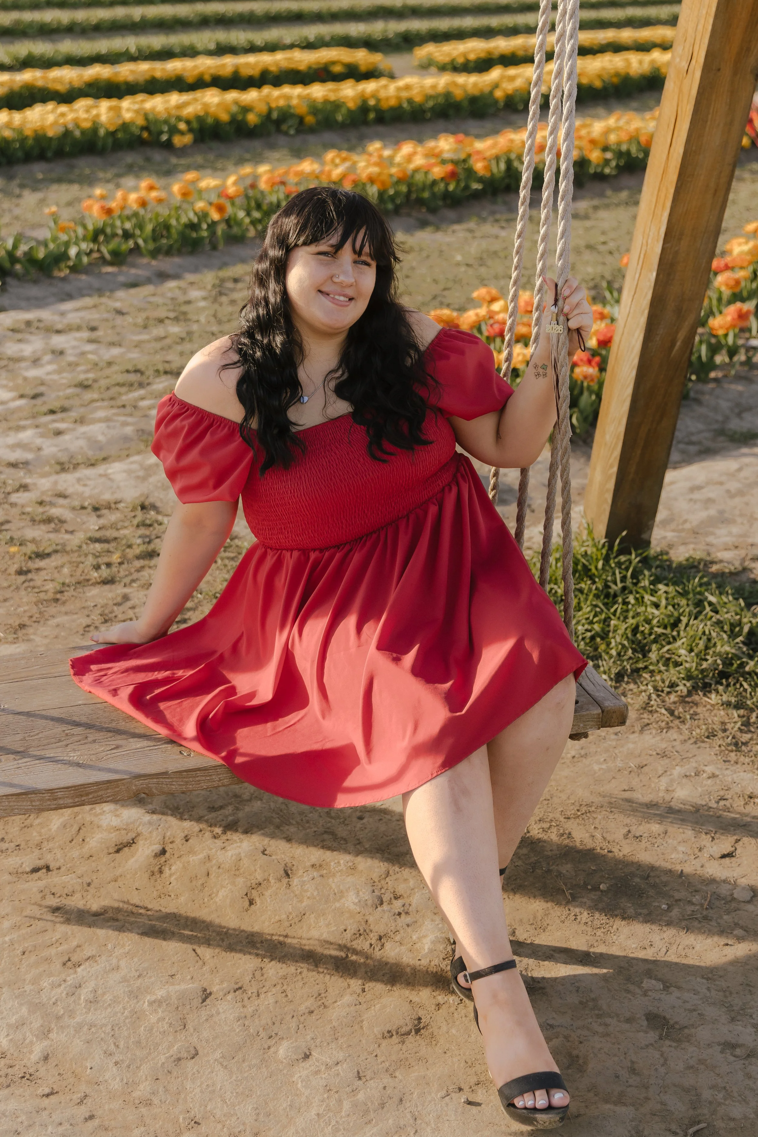 A woman sitting on a wooden swing in a flower field, wearing a red dress and black heels, smiling.