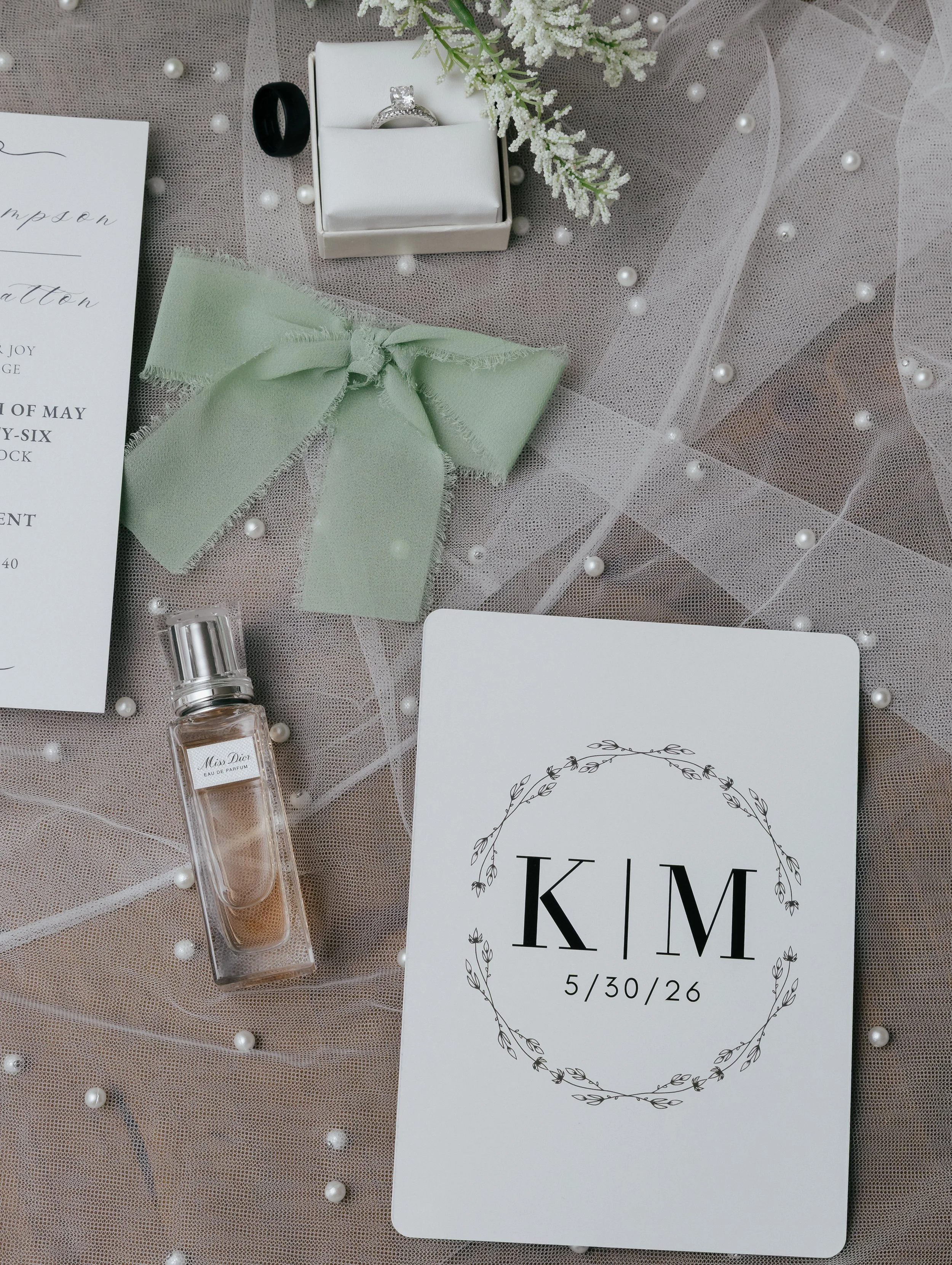 Wedding invitation with initials K & M and date 5/30/26, surrounded by pearl beads, green bow, perfume bottle, engagement ring in a box, and decorative white flowers on mesh fabric.