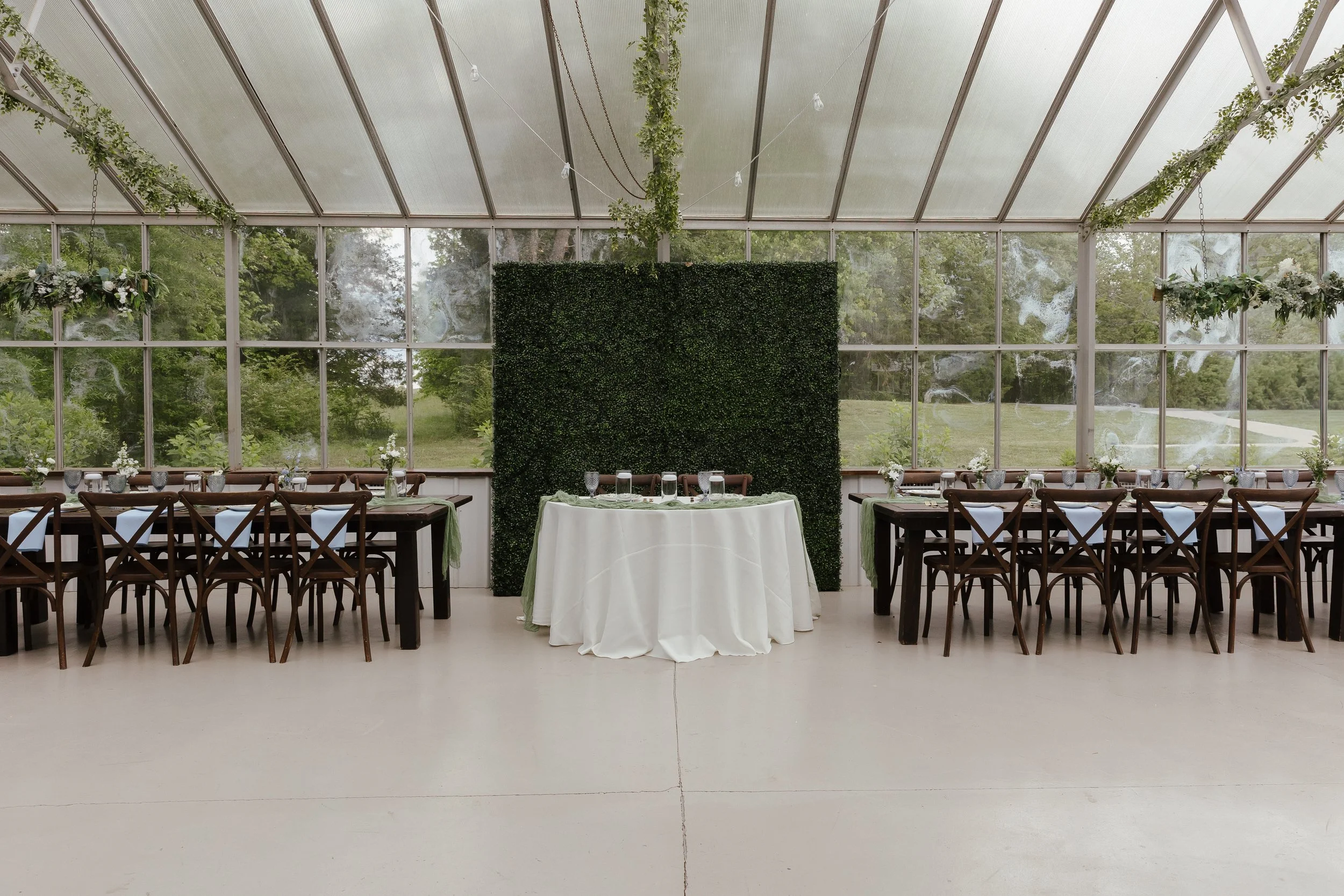 A photo of a wedding reception space in a green house with a greenery backdrop, blue napkins, glasses, and long wooden tables.