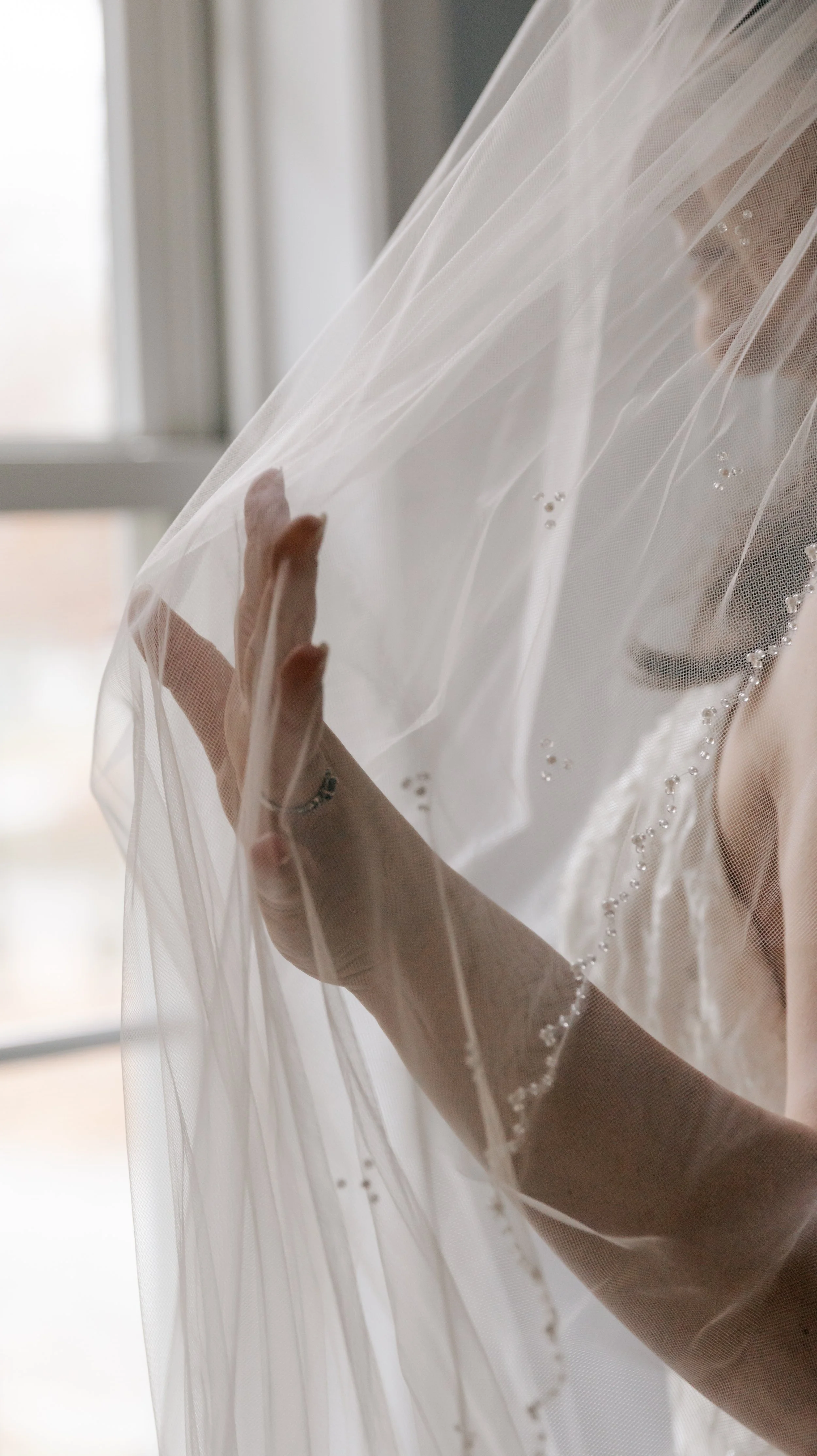 Close-up of a bride’s hand touching a sheer wedding veil near a window with natural light.
