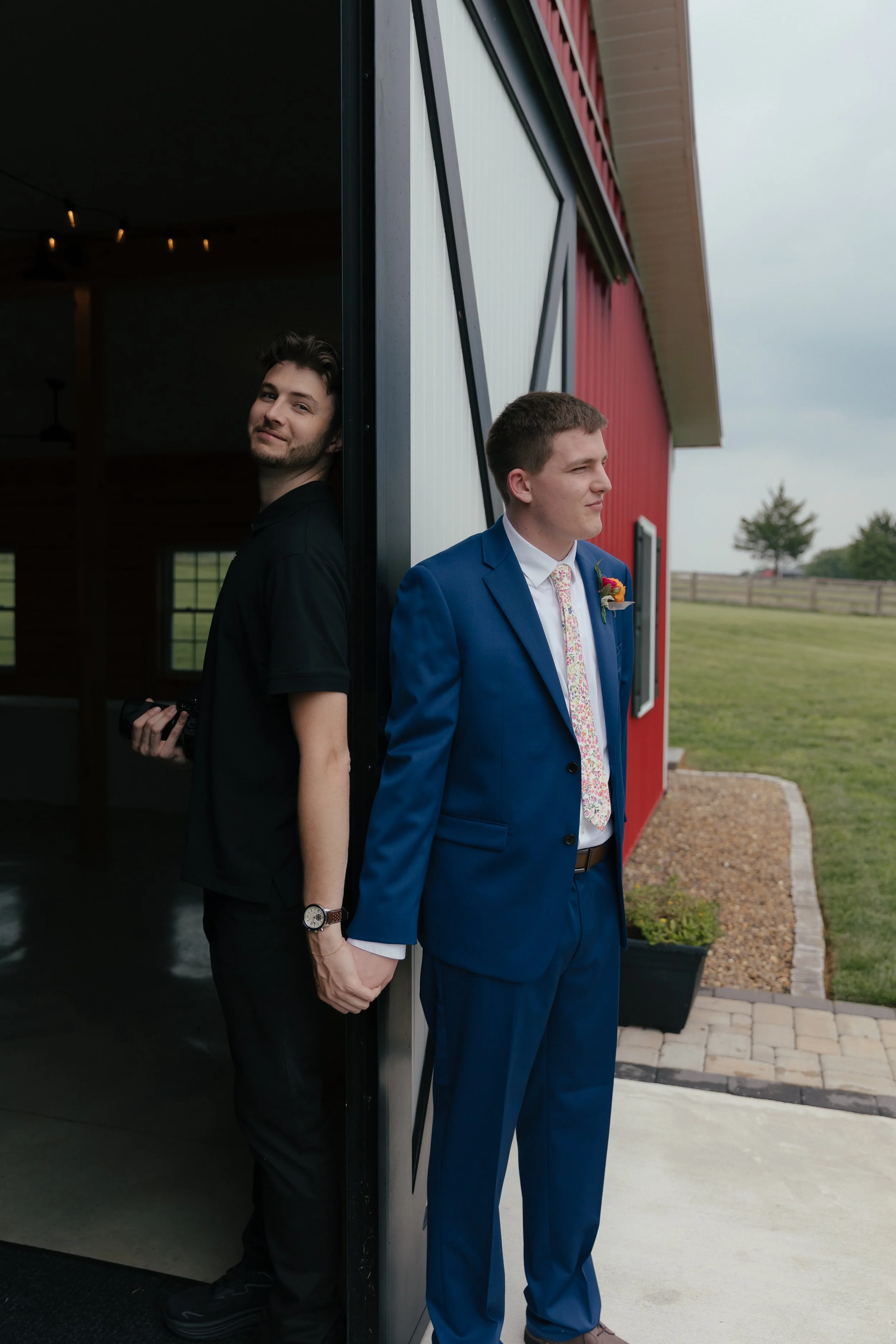 A man holding a camera on one side of a barn door, holding the hand of a groom for a funny photo. with the groom before a first touch and prayer.