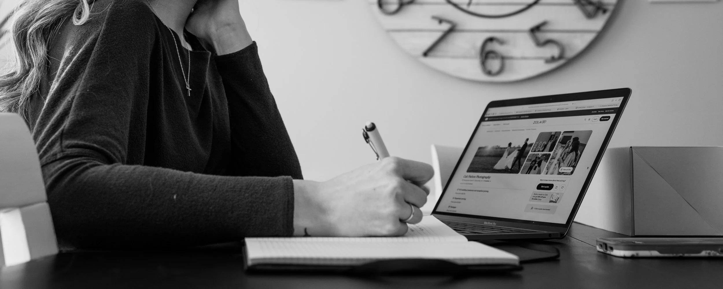 A woman in a long sleeve shirt sitting at a desk, looking at a laptop screen with a photography website, holding a pen, with a notebook in front of her, and a clock hanging on the wall behind.