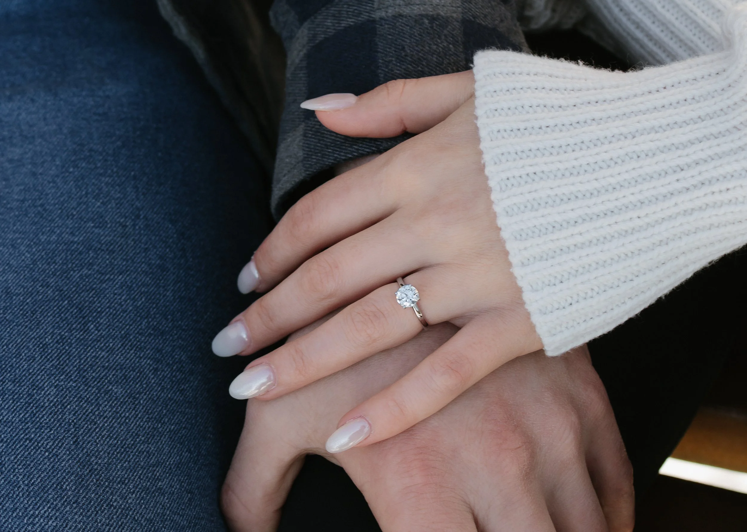 Close-up of a woman's hand with a large diamond engagement ring on her ring finger, resting on a man's hand. The woman is wearing a white knitted sweater, and the man is dressed in dark clothing, likely jeans.