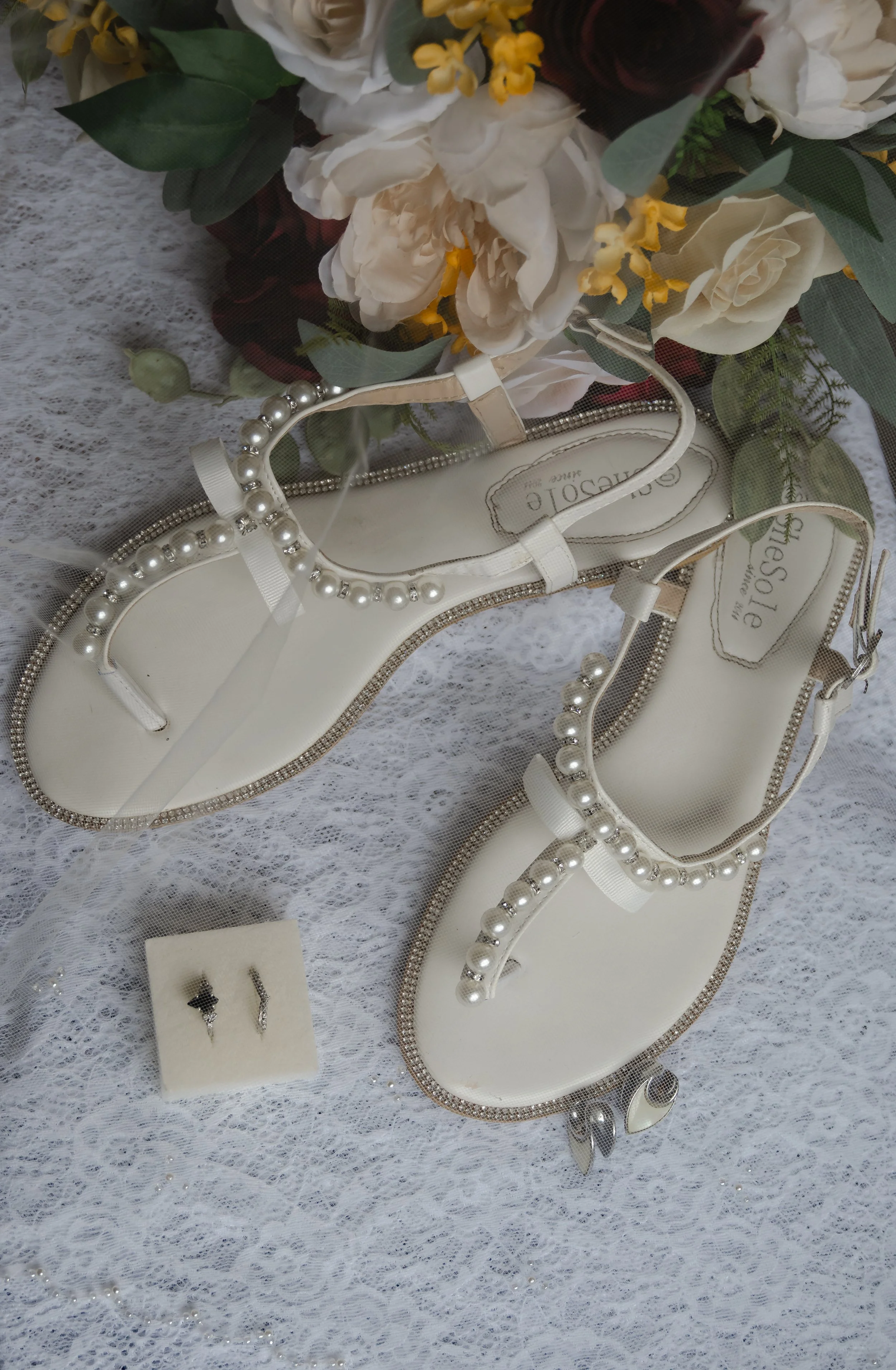 A pair of white sandals decorated with pearls, placed on lace fabric, alongside a small jewelry box with earrings, and a bouquet of white and yellow flowers.