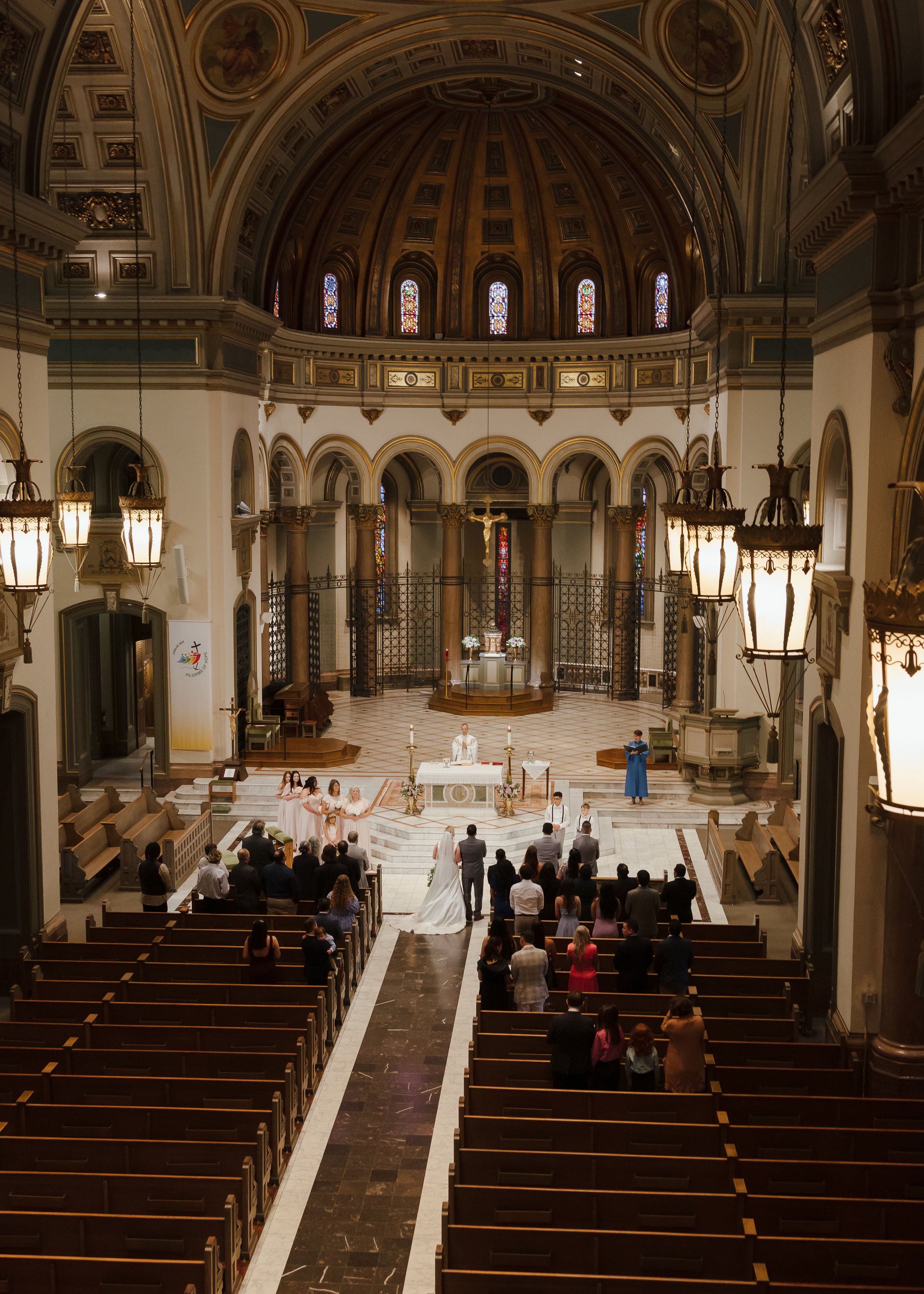 A wedding ceremony taking place inside a grand church with a high domed ceiling. The altar is decorated with flowers, and a priest is officiating. The bride and groom are standing at the front, surrounded by wedding attendants and guests seated in wo