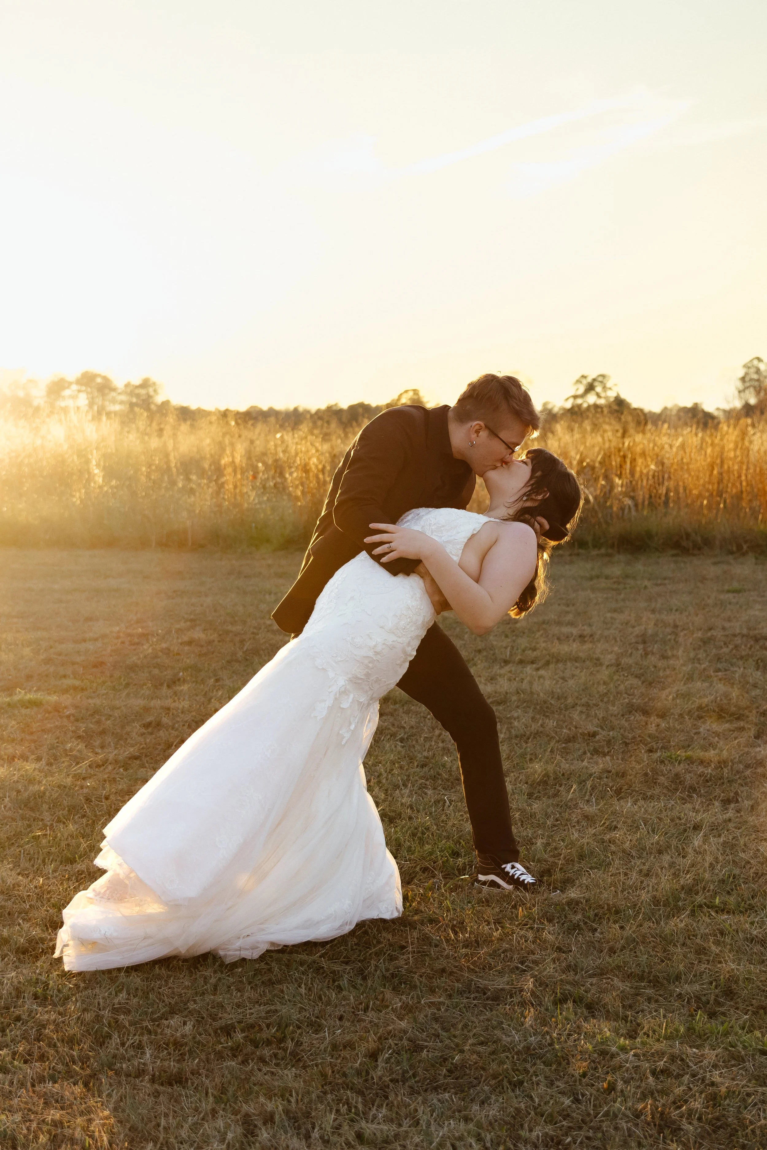 A couple in wedding attire sharing a kiss in a field at sunset, with the groom dipping the bride backwards.