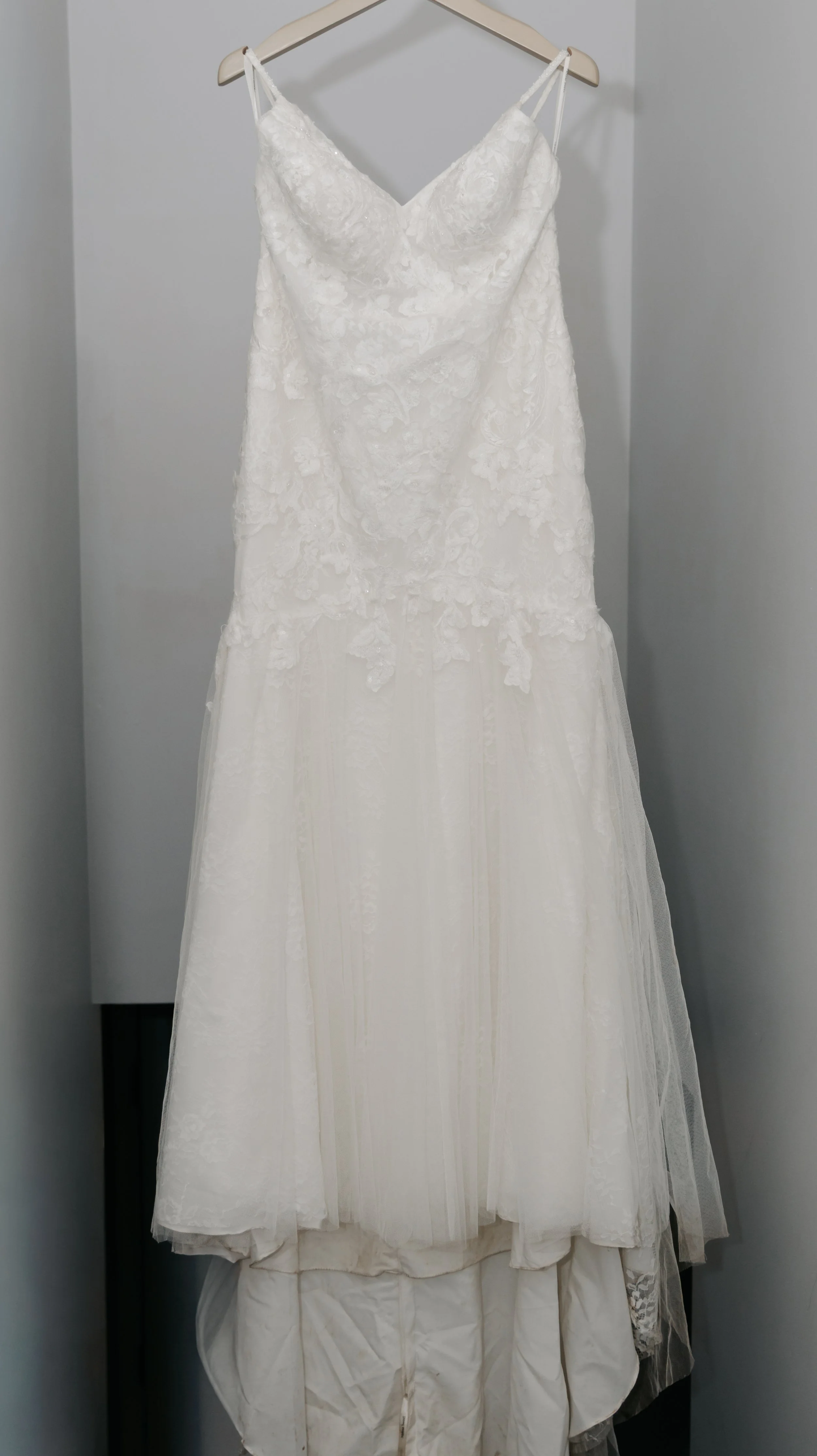 A white wedding dress with lace details on a hanger against a gray wall.
