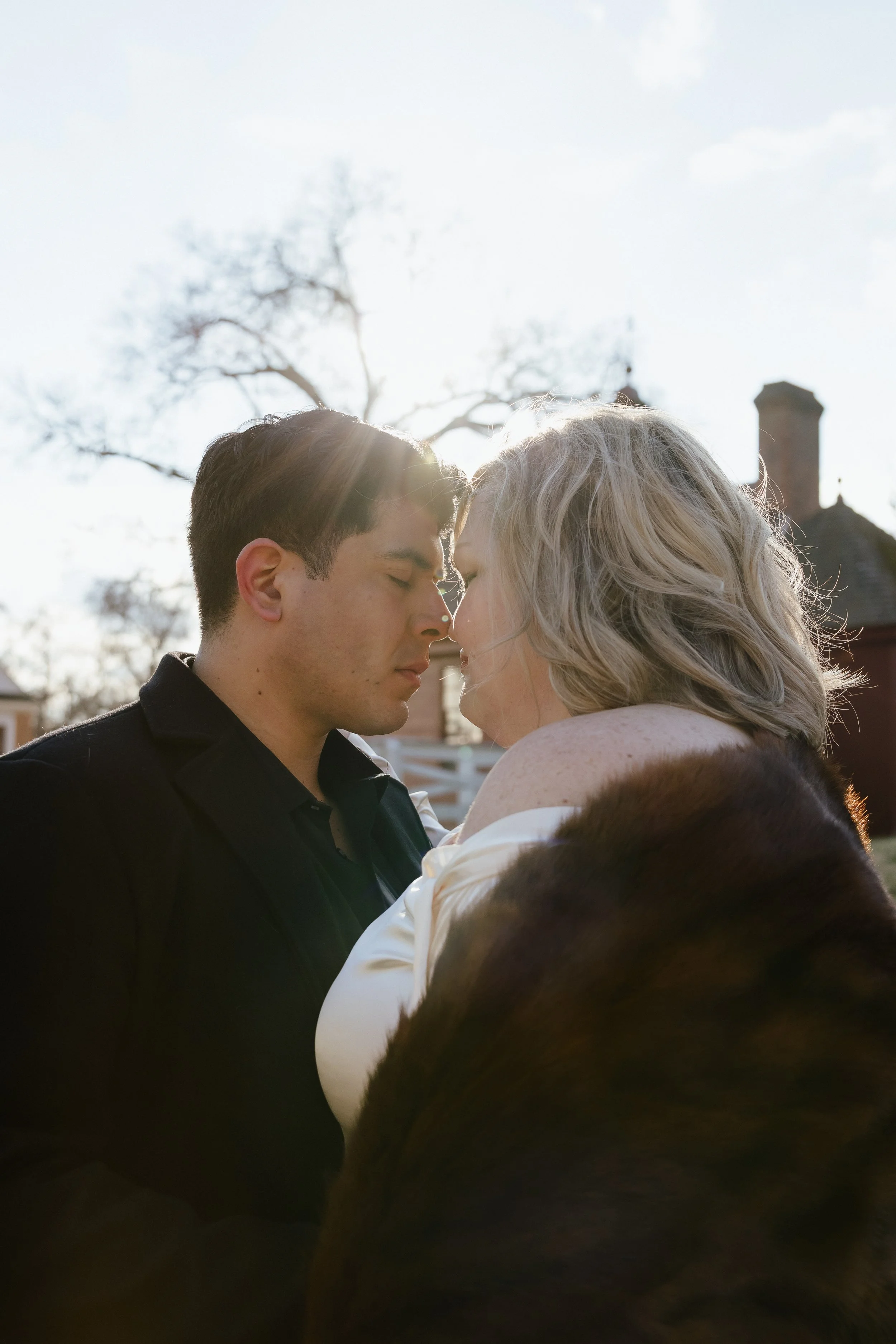 A close-up of a couple with their foreheads touching, standing outdoors during sunset, with trees and houses in the background.