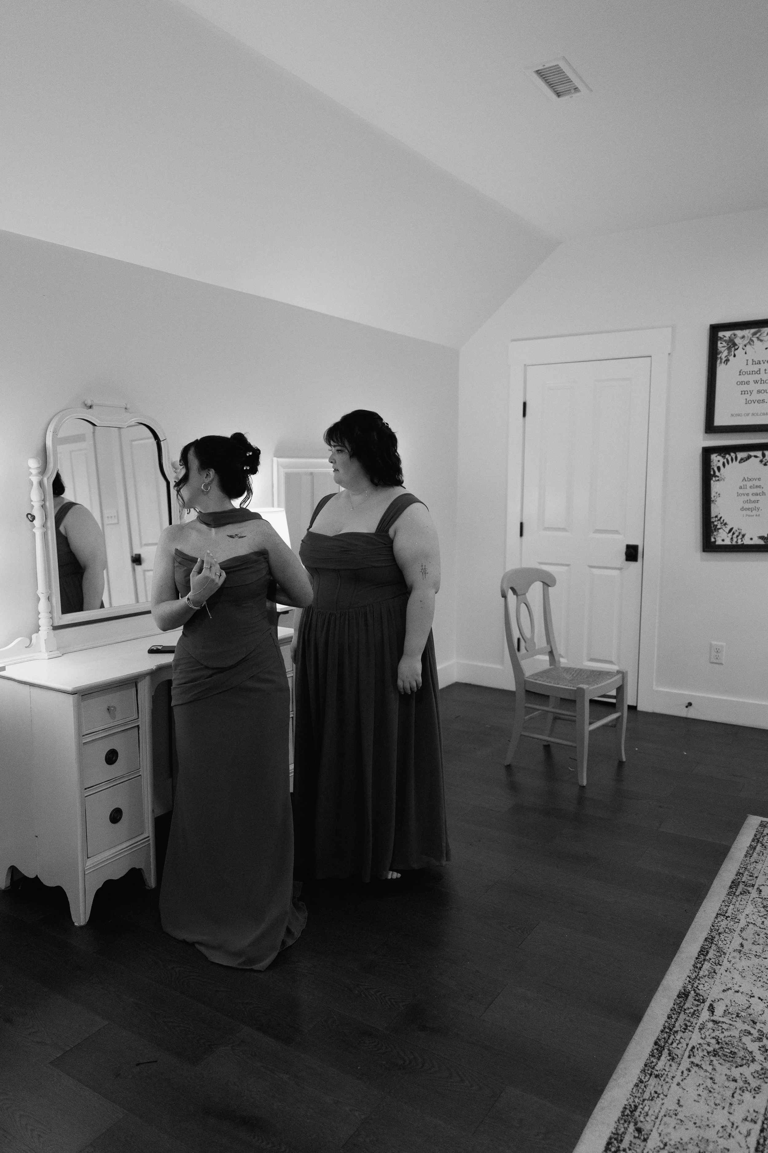 A black and white photo of two girls looking at their dresses in a vanity mirror. 