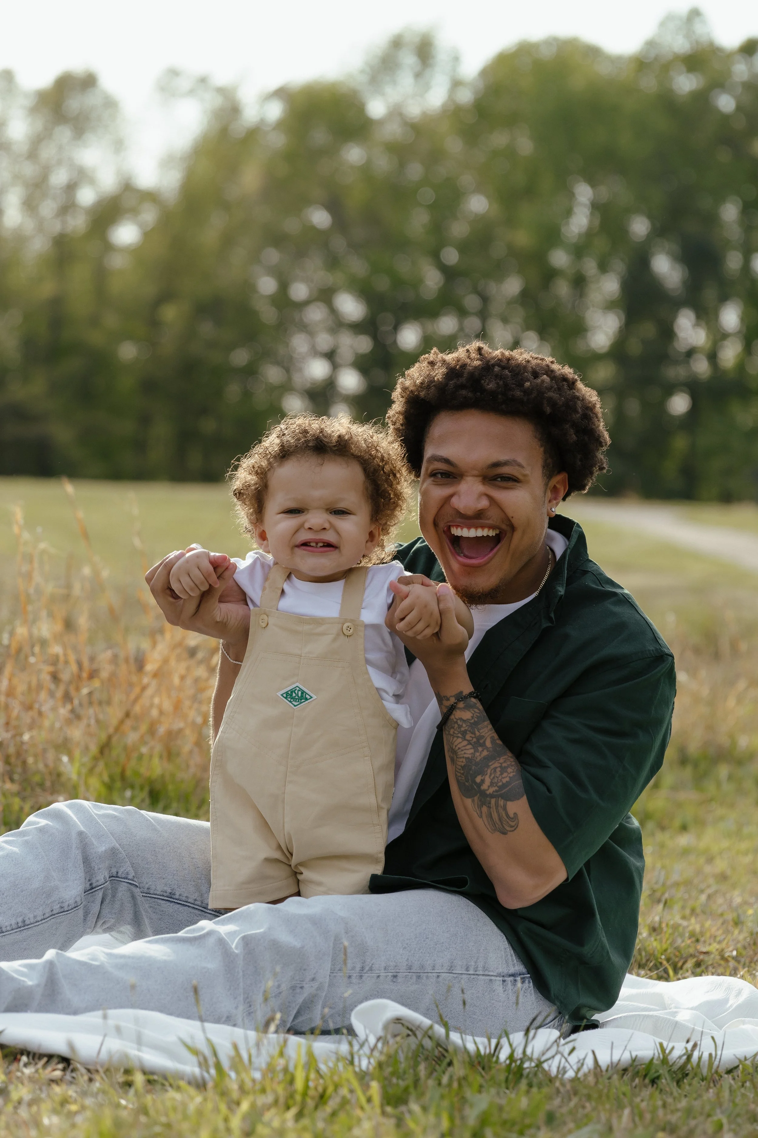 A man and a young child sitting on a blanket outdoors in a grassy field, smiling and making faces at the camera.