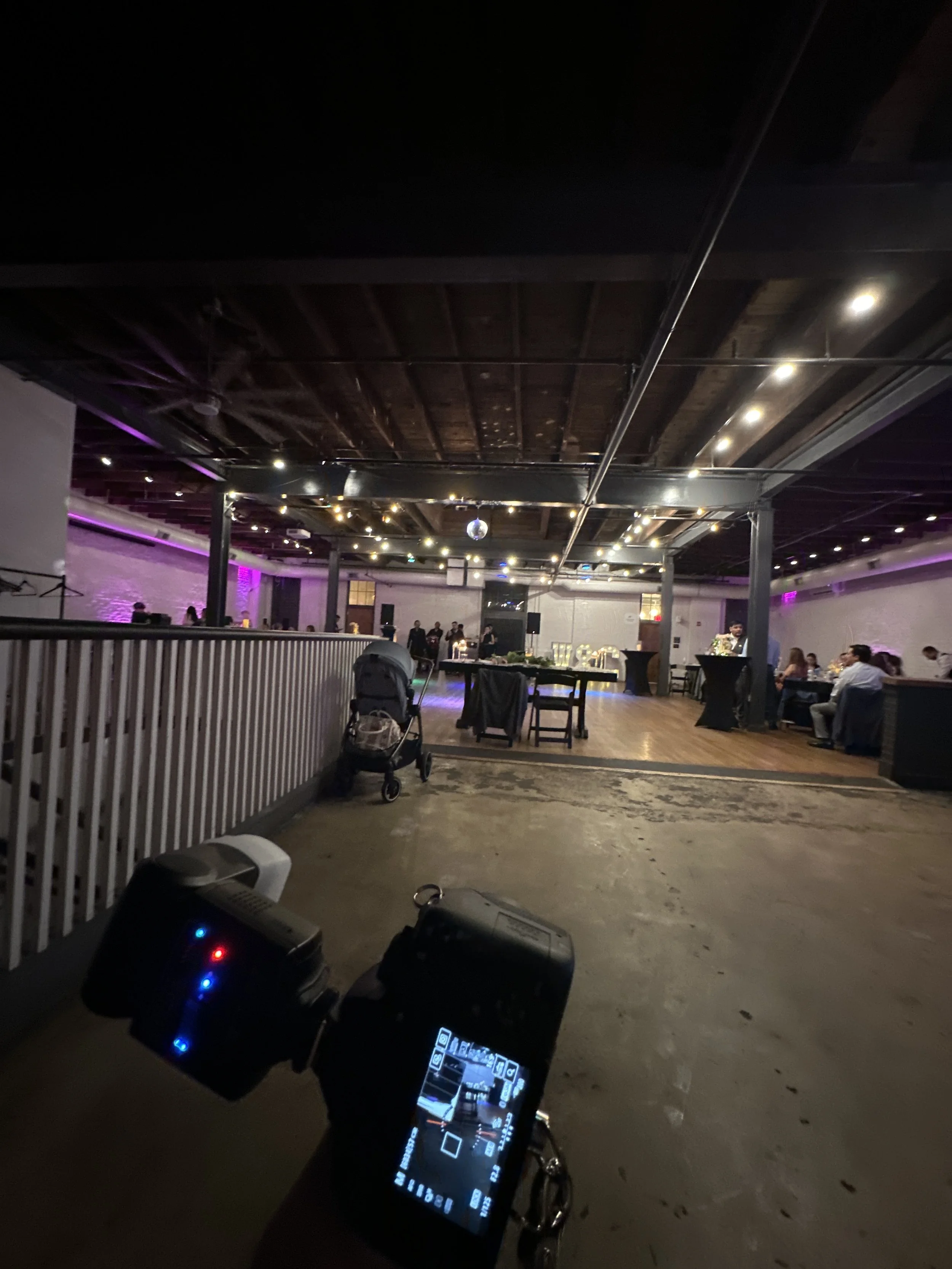 Empty indoor event space with tables, chairs, some people, and purple lighting, taken from a camera held by a person.