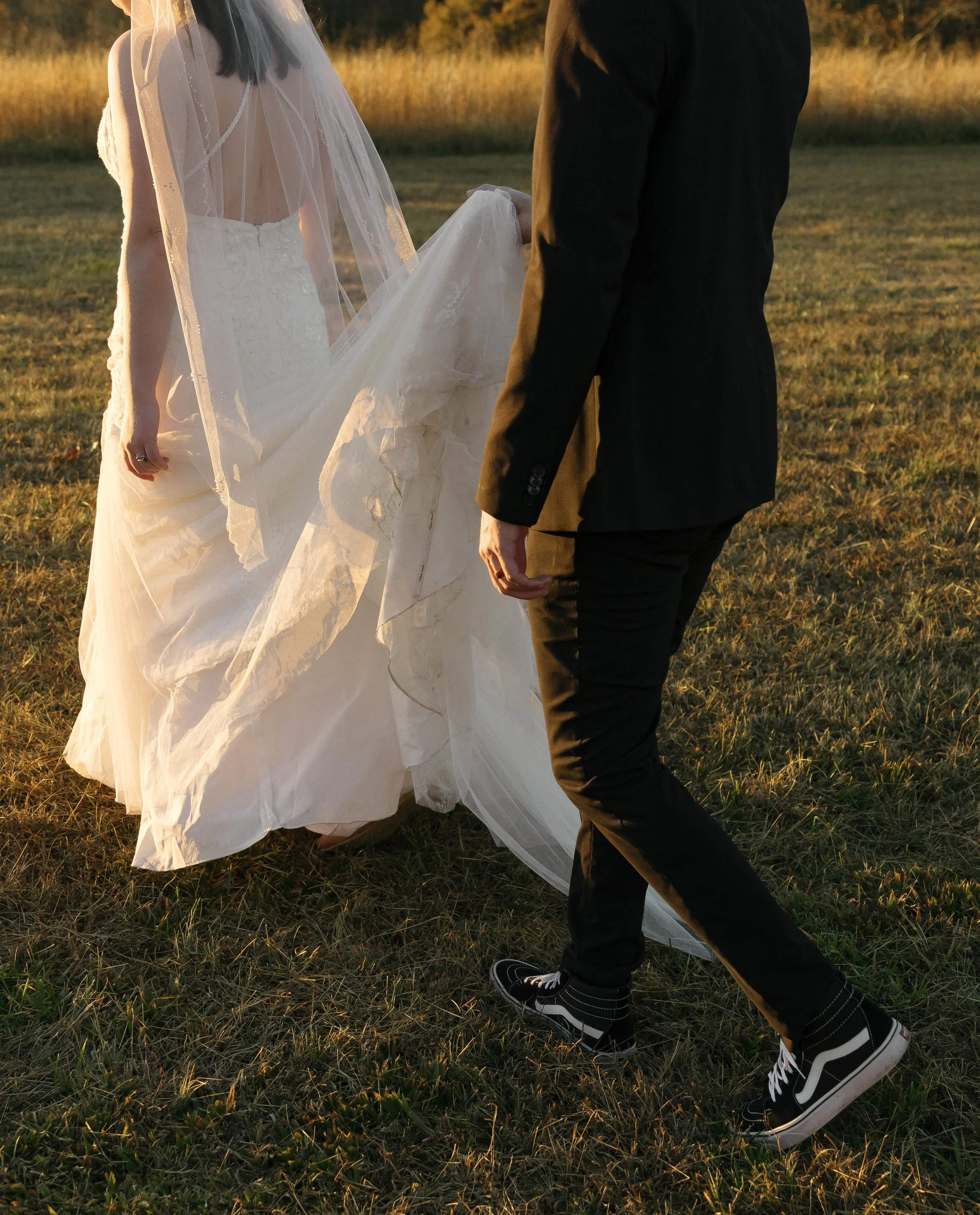 A bride and groom walking outdoors during sunset, with the bride holding up her wedding dress as they walk on a grassy field.