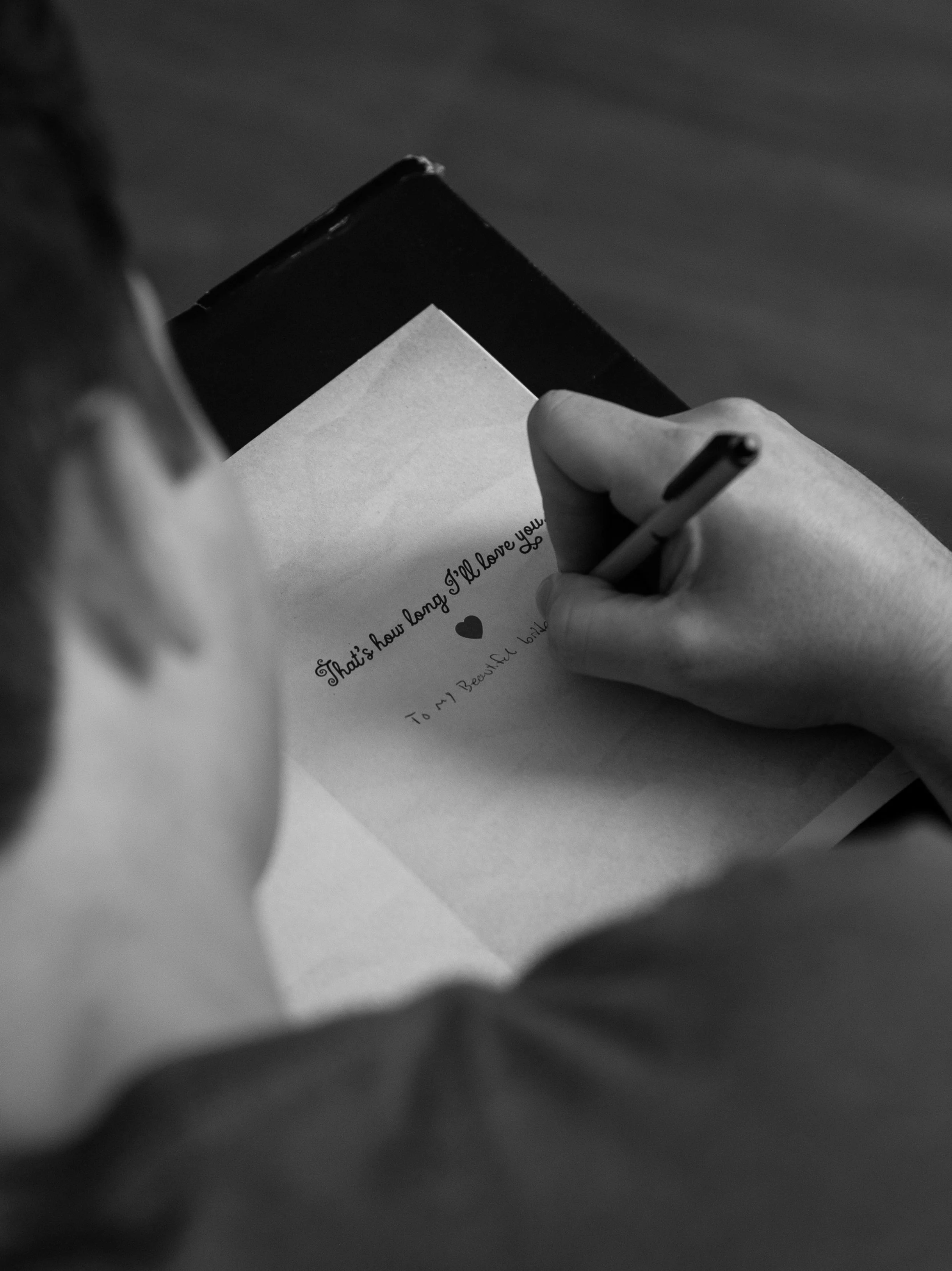 A black and white photo of an over the shoulder perspective of the groom writing in the card he will be giving to his soon to be wife.