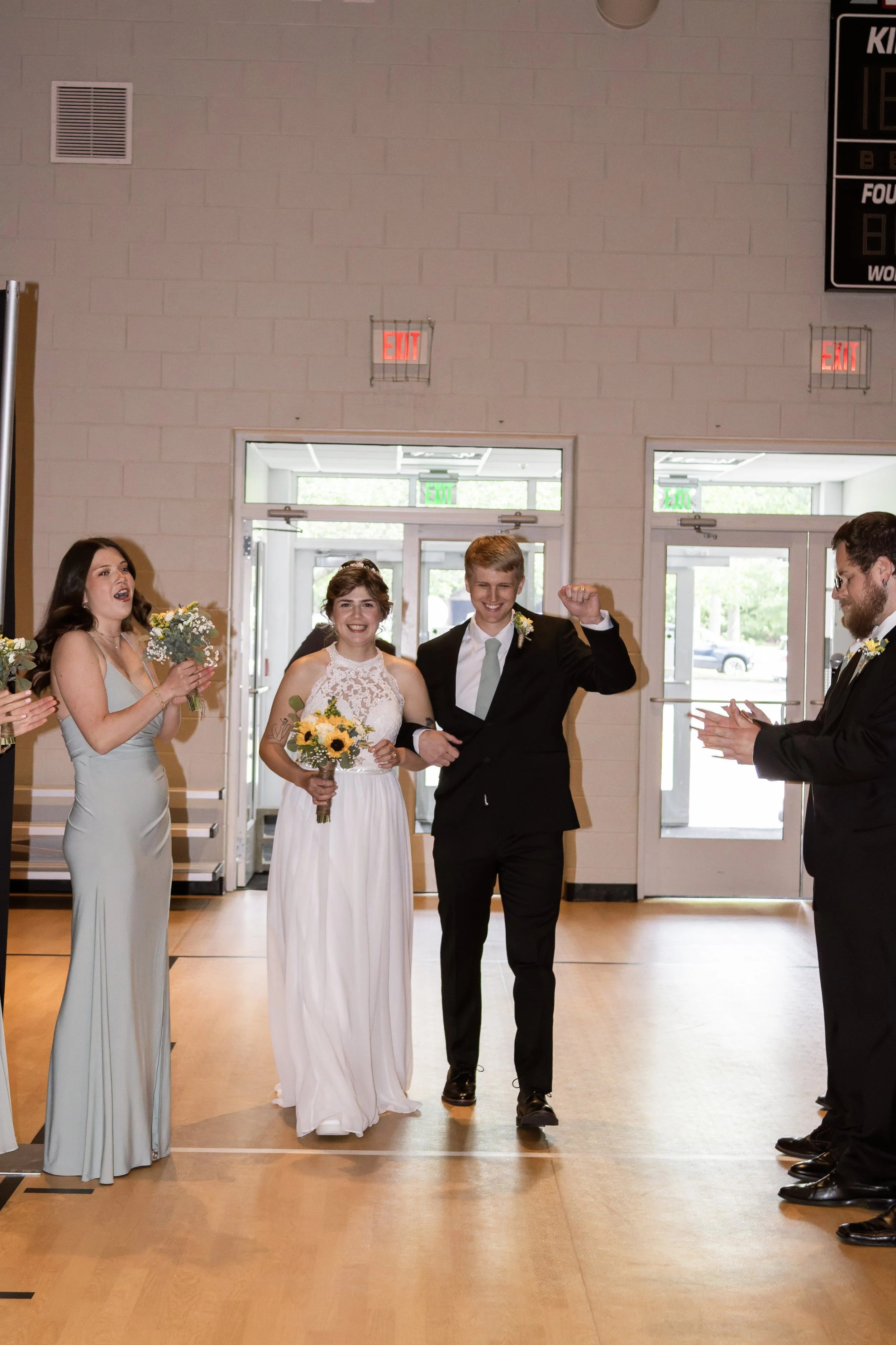 A wedding celebration with a bride and groom walking into the room, surrounded by friends clapping and smiling.