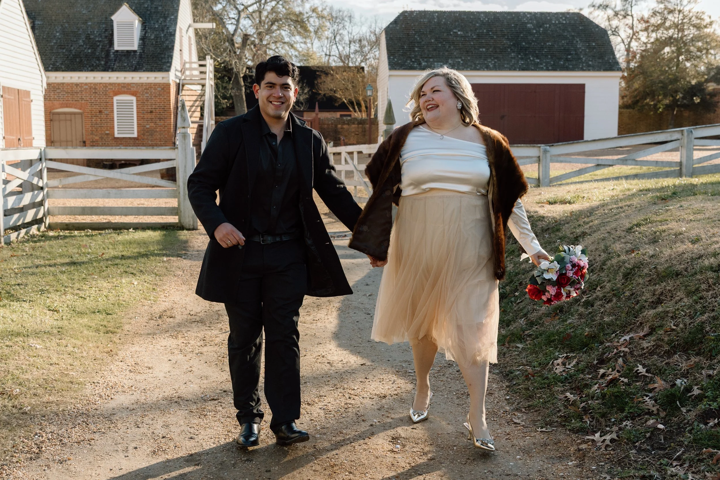 A smiling man and woman holding hands and walking on a dirt path outdoors during daytime, with the woman holding a bouquet of flowers. The woman is dressed in a cream-colored skirt with a satin top and a brown fur stole, while the man is wearing a bl