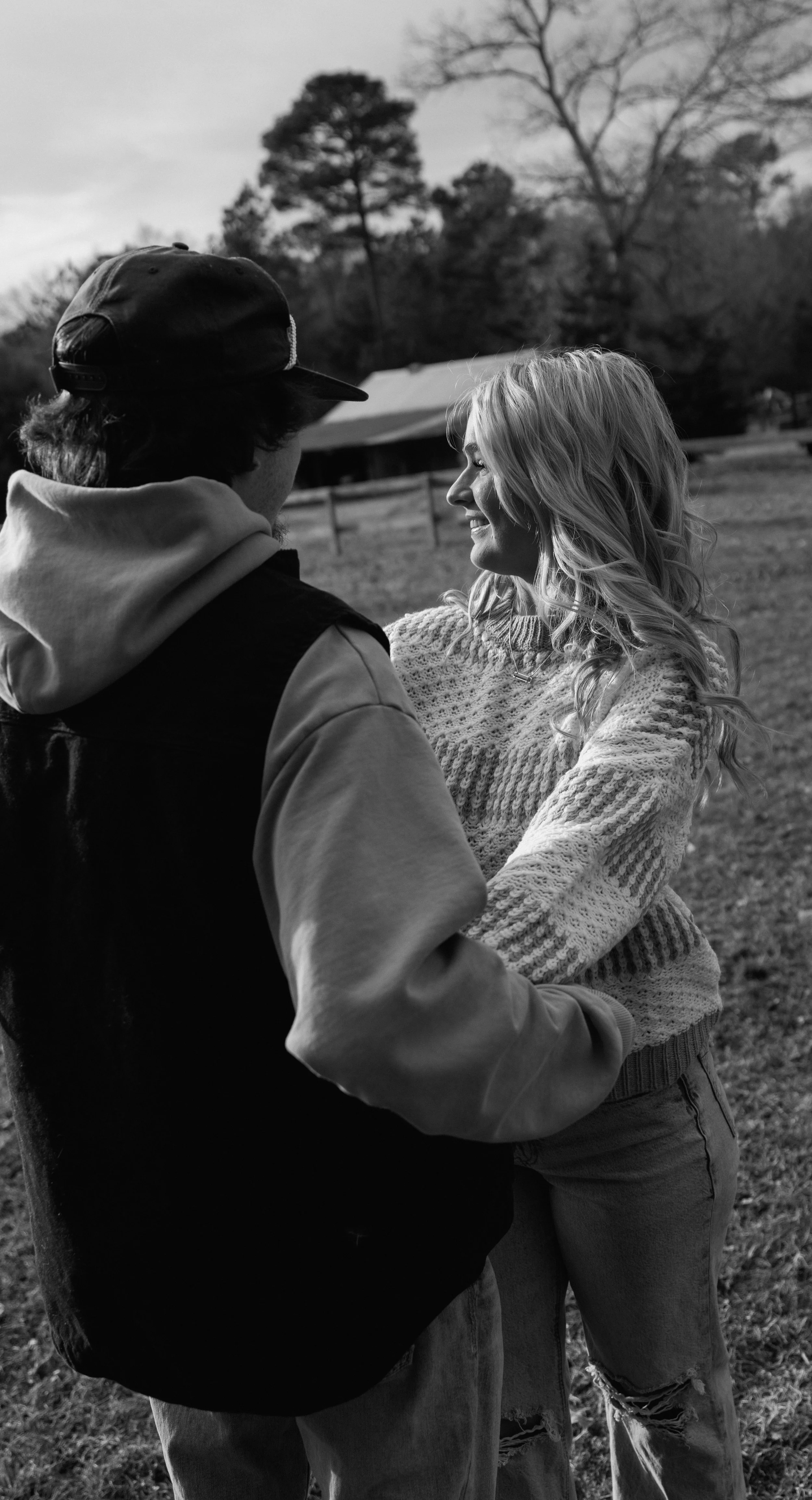 A black-and-white photo of a man and woman smiling and looking at each other outdoors on a cloudy day, with trees and a building in the background.