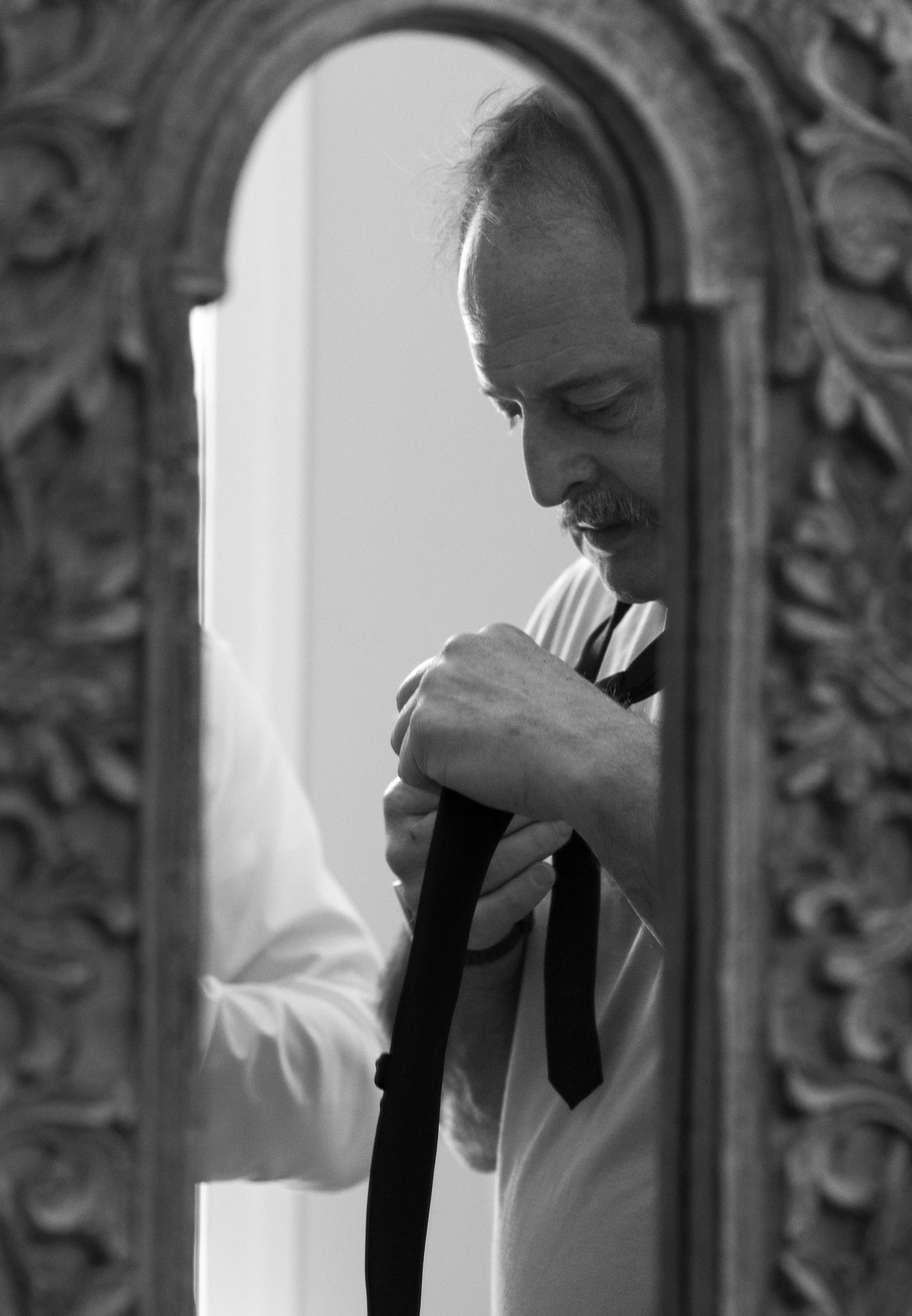 A black and white mirror reflection photo of a groom's father tying the groom's tie for him on his wedding day. The mirror is small and has ornate floral detail carvings surrounding the mirror.