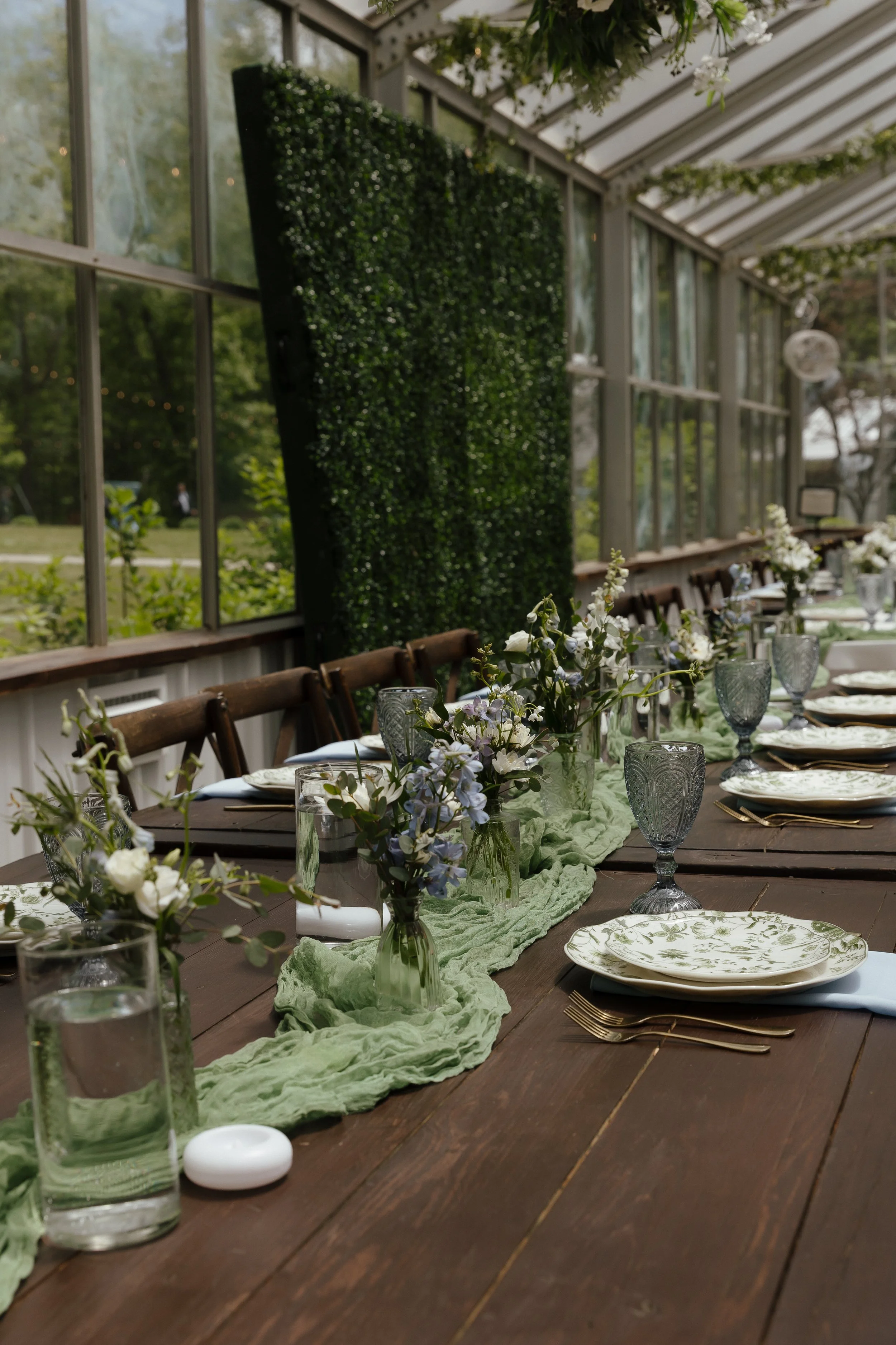 A photo of a reception table's details in a greenhouse with greenery backdrops, small florals in vases, a sage green cheesecloth runner, and blue goblet glasses. The plates have an intricate green floral design on a white background.
