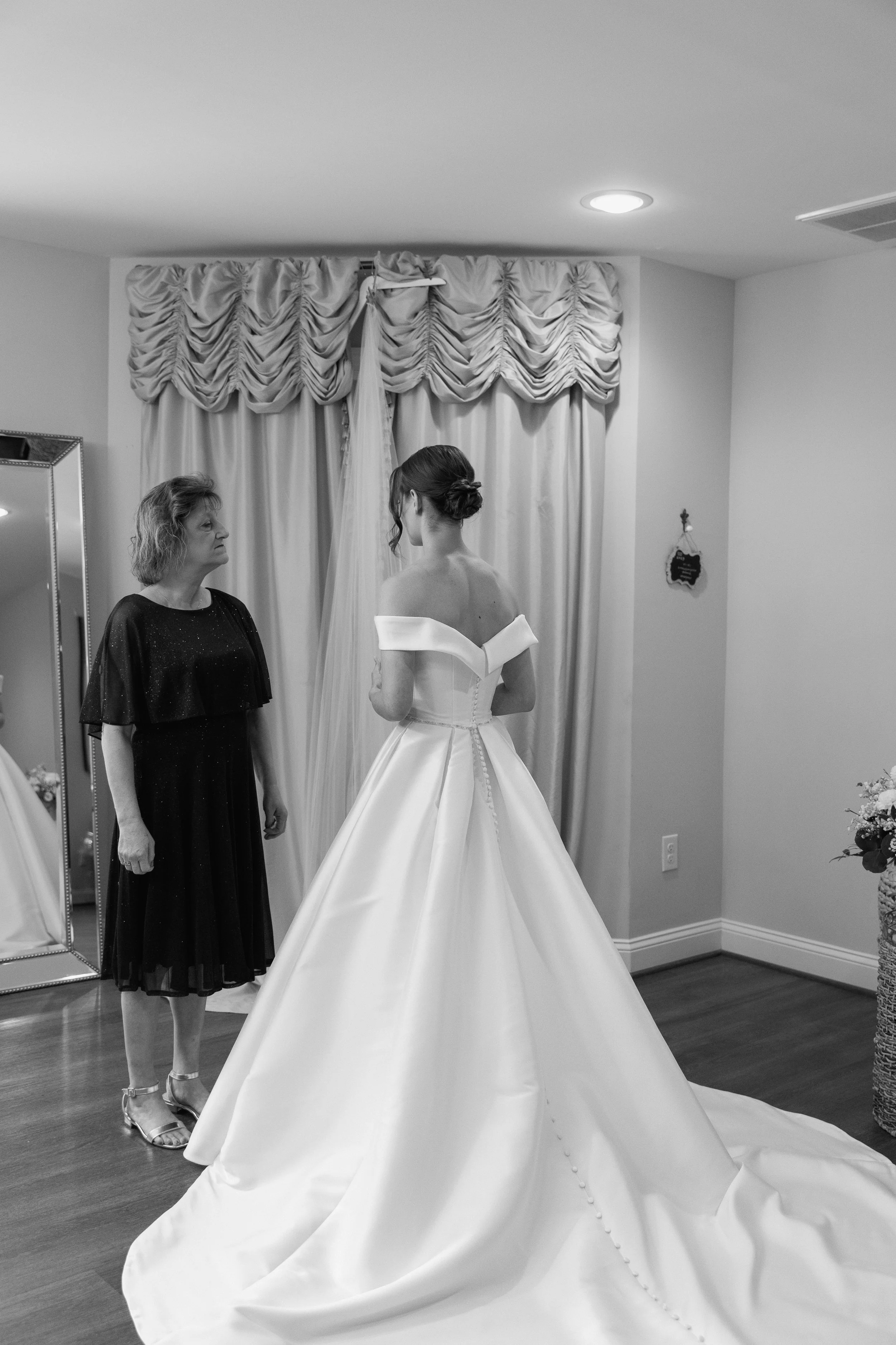 A bride in an elegant strapless wedding gown with a long train stands facing an older woman in a dark dress, in a room with draped curtains and a mirror.