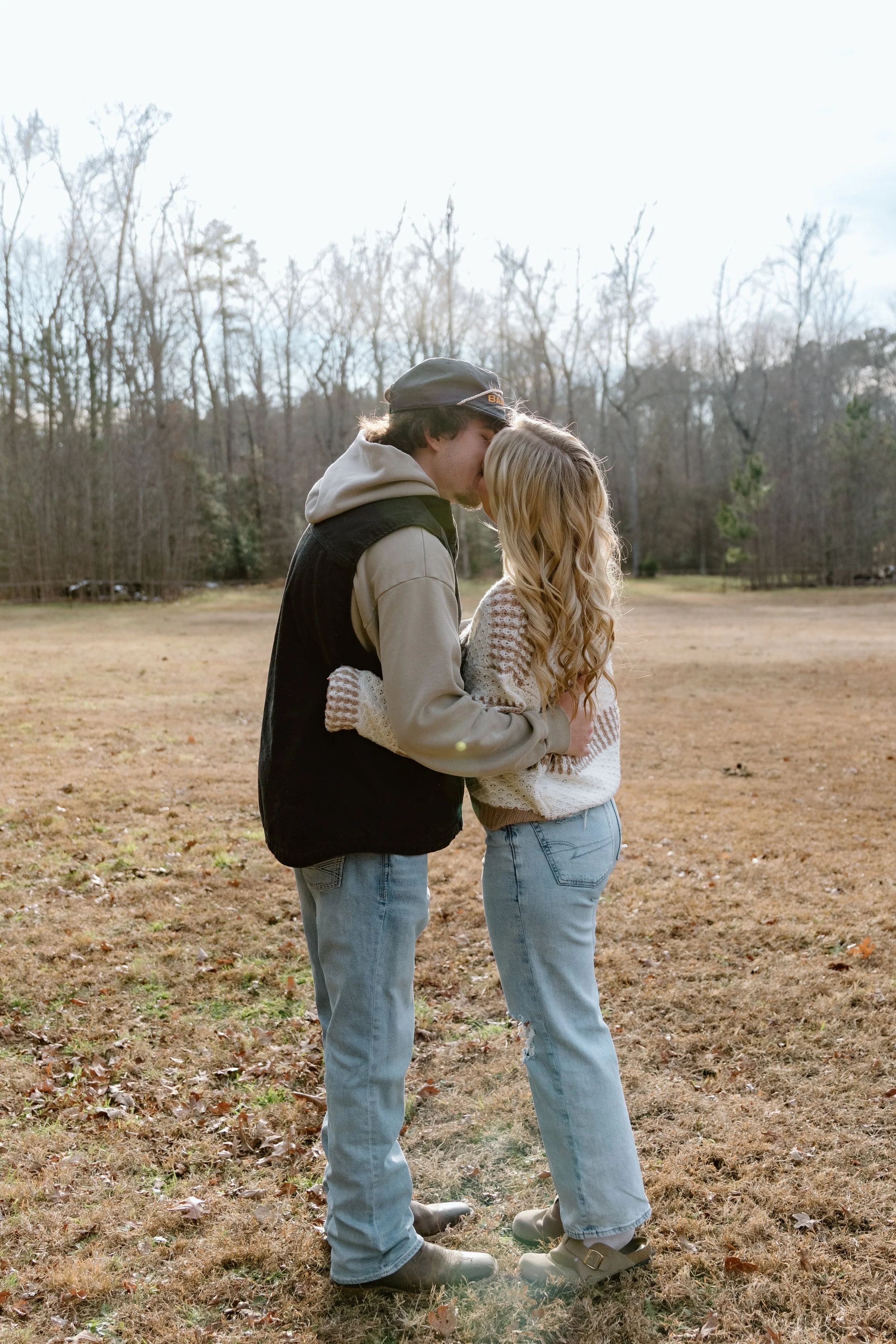 A young couple kissing outdoors in a field, with bare trees in the background during late fall or winter.