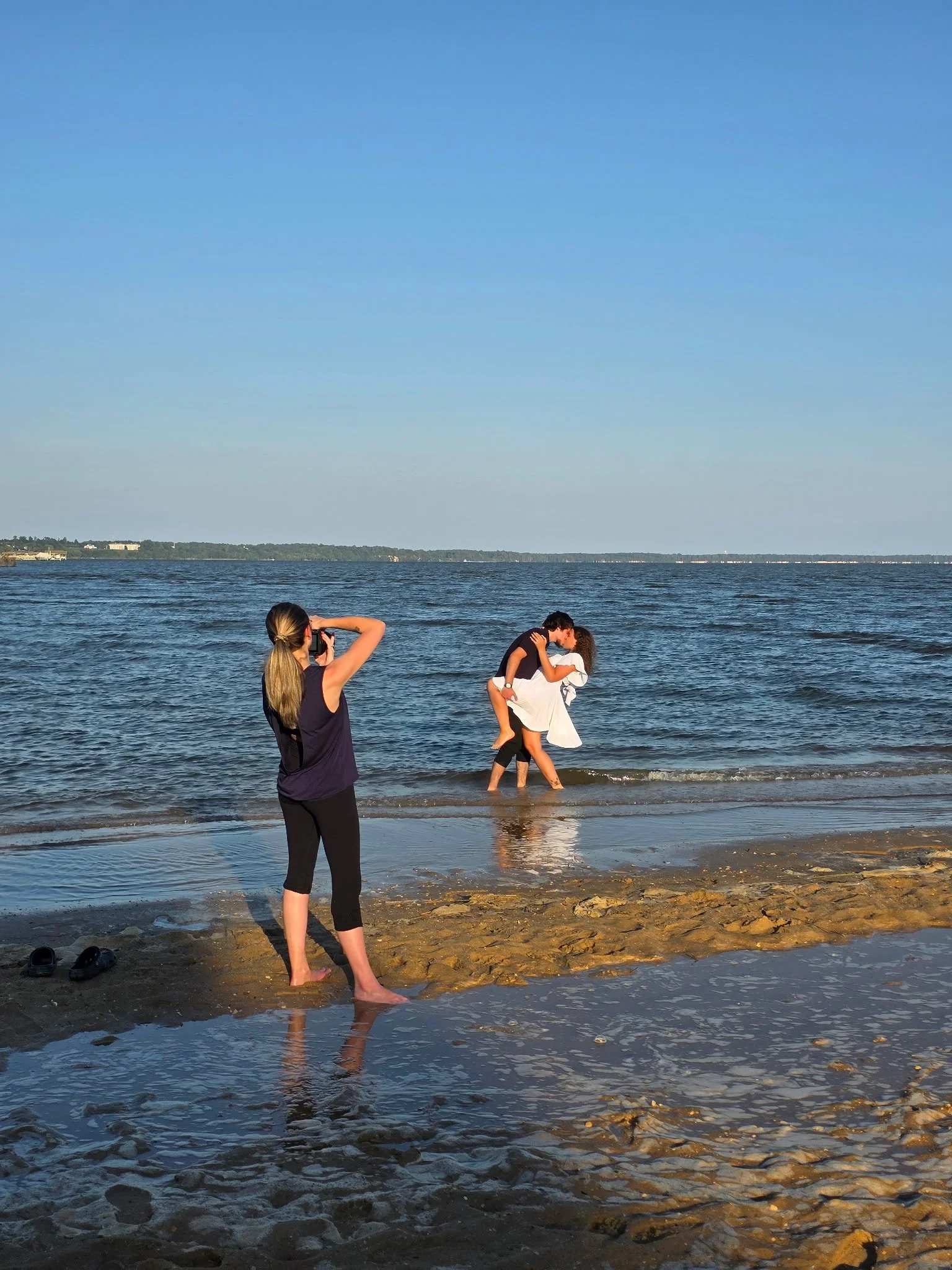 A woman stands on a beach taking photographs of a couple embracing in the water. The man lifts the woman while they kiss near the shoreline, with a clear blue sky above.