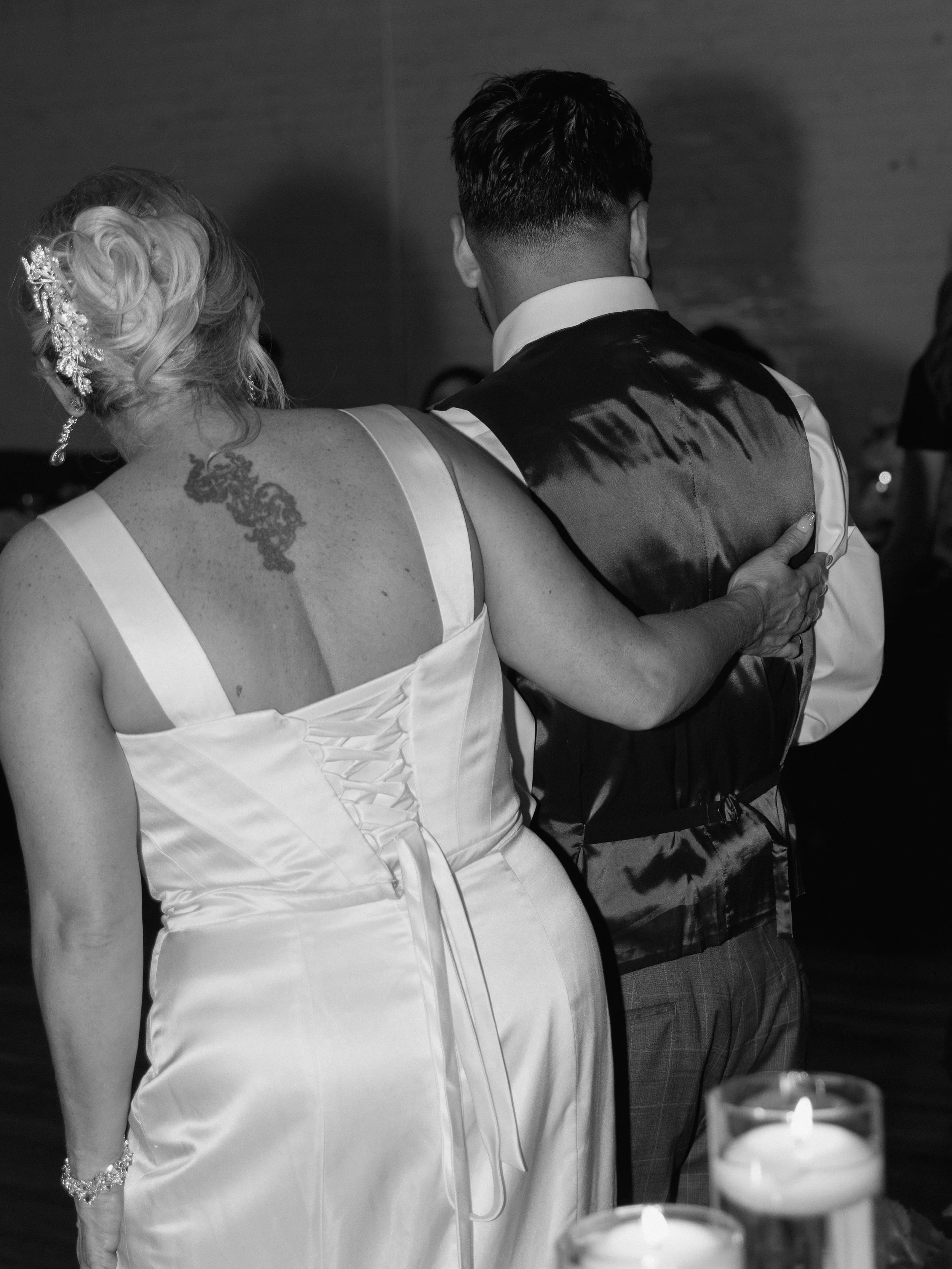 Black and white photo of a couple dancing at a formal event, the woman has blonde hair styled in curls with an ornate hairpiece and a tattoo on her shoulder, and is wearing a white dress. The man has dark hair and is wearing a tuxedo with a satin bac