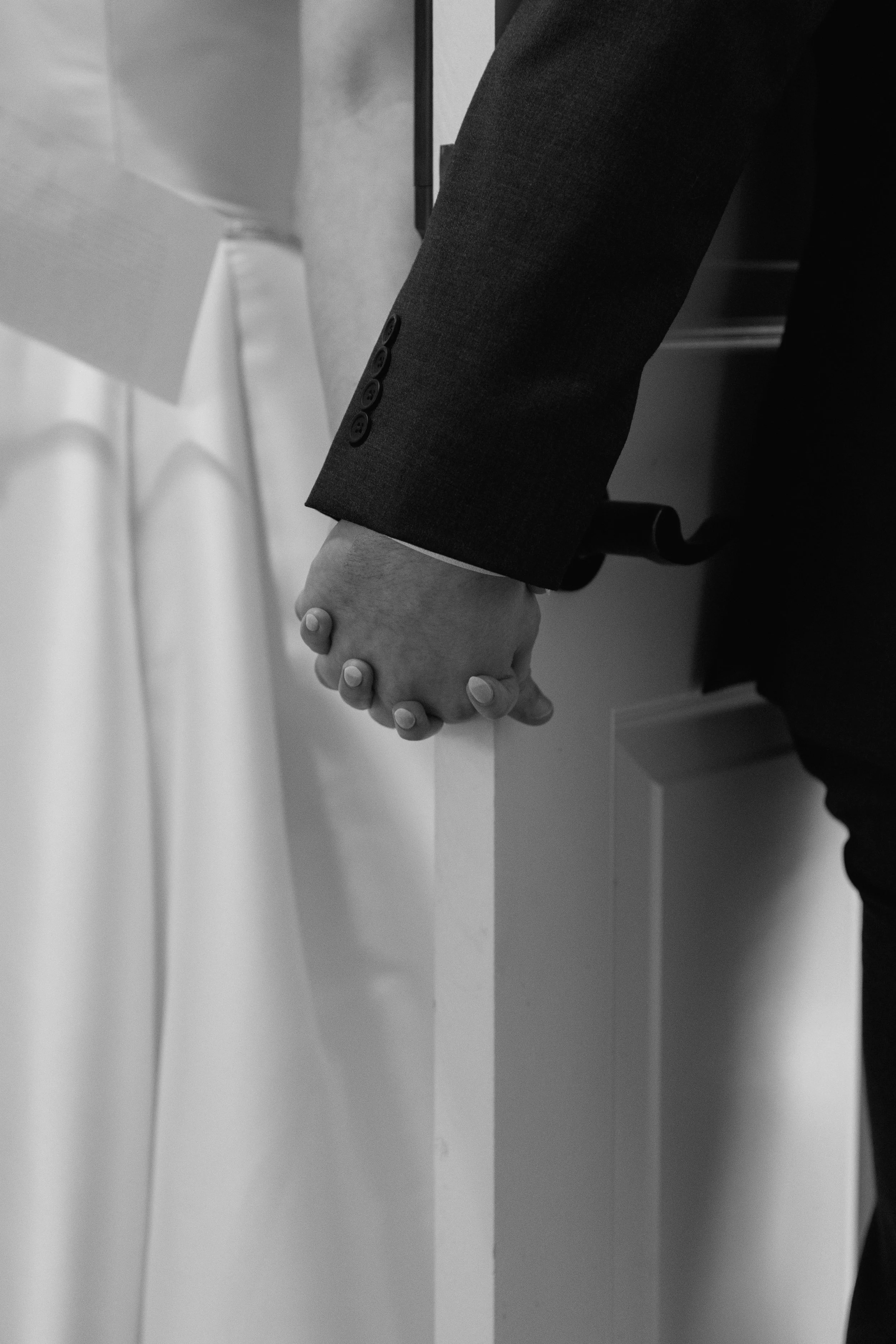 A person in a dark suit with a buttoned sleeve is holding hands with another person in a white attire, standing in front of a white surface or wall.