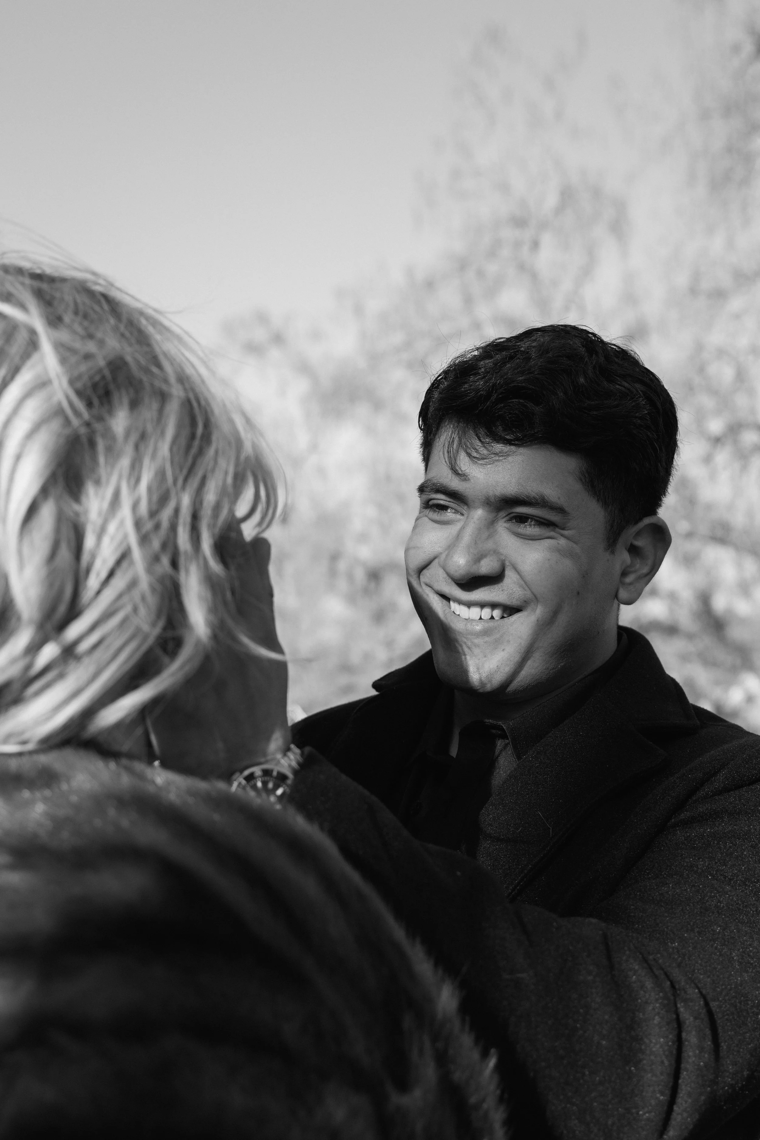 A black-and-white photo of a young man smiling and looking at a woman with blonde hair, outdoors with a blurred background.