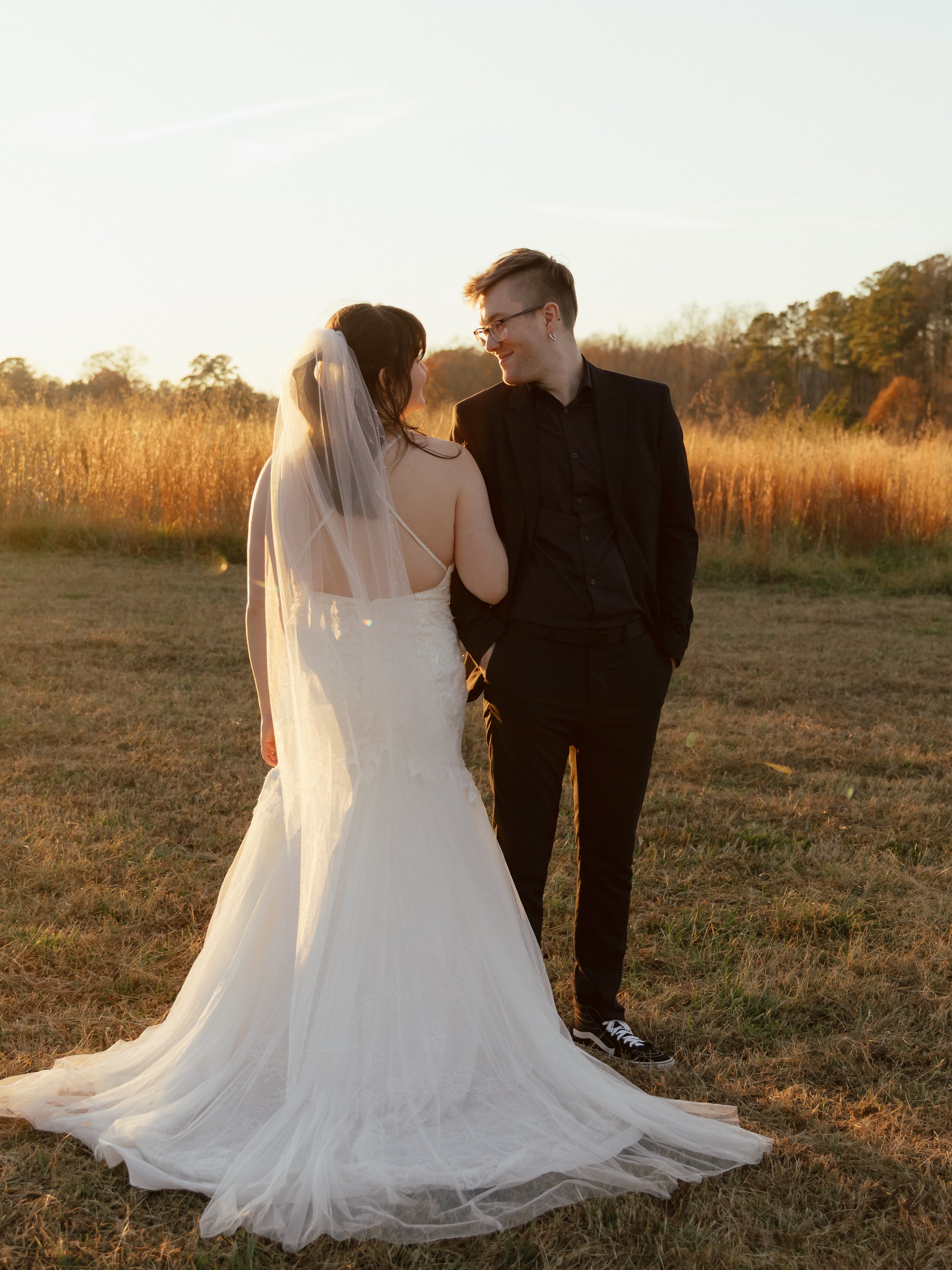 A bride and groom standing close together outdoors during sunset, with the bride in a white wedding gown and veil, and the groom in a black suit, smiling at each other in a field of tall grass.