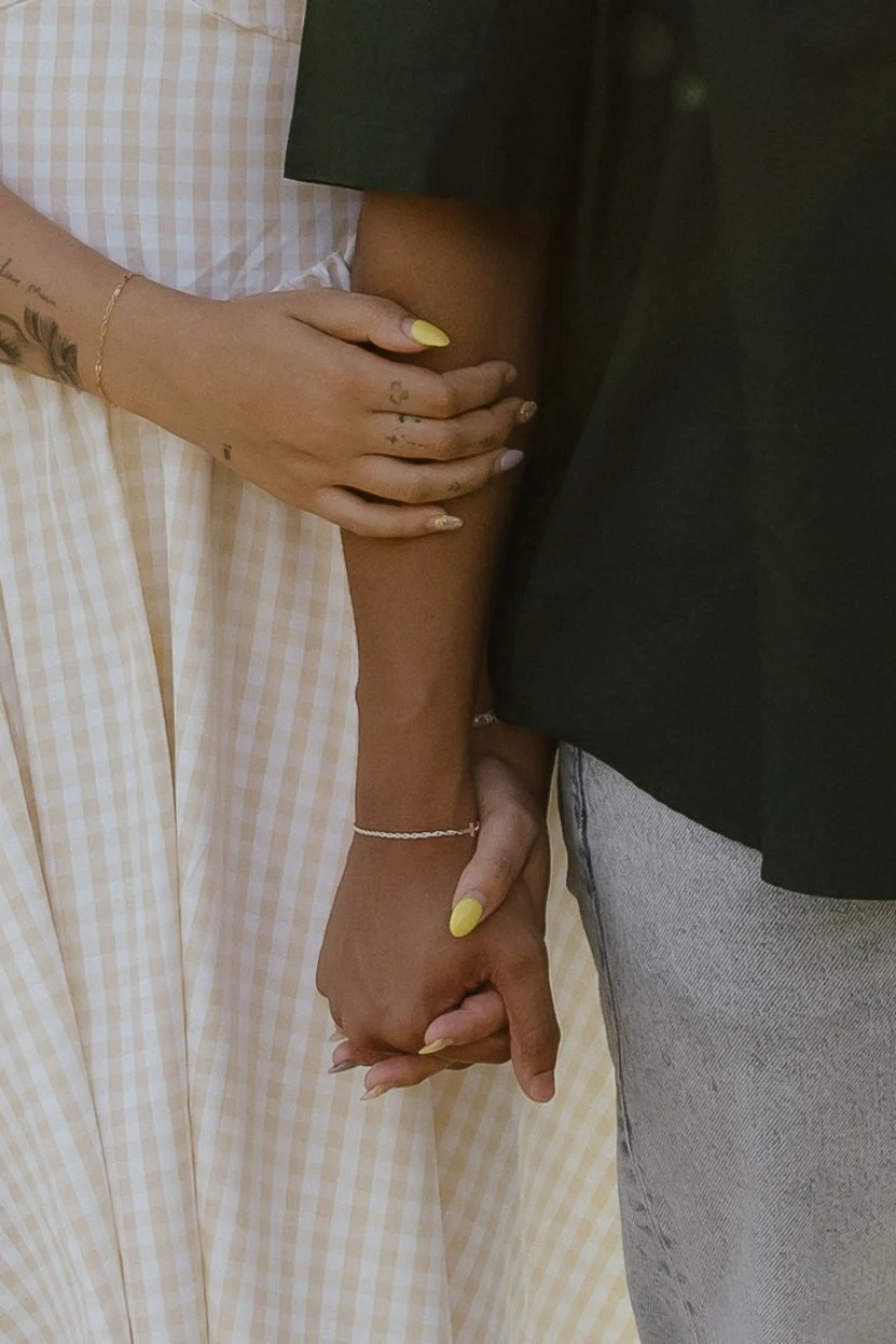 Two people holding hands, one with painted nails and a tattoo, the other with a bracelet, in front of a checkered fabric background.