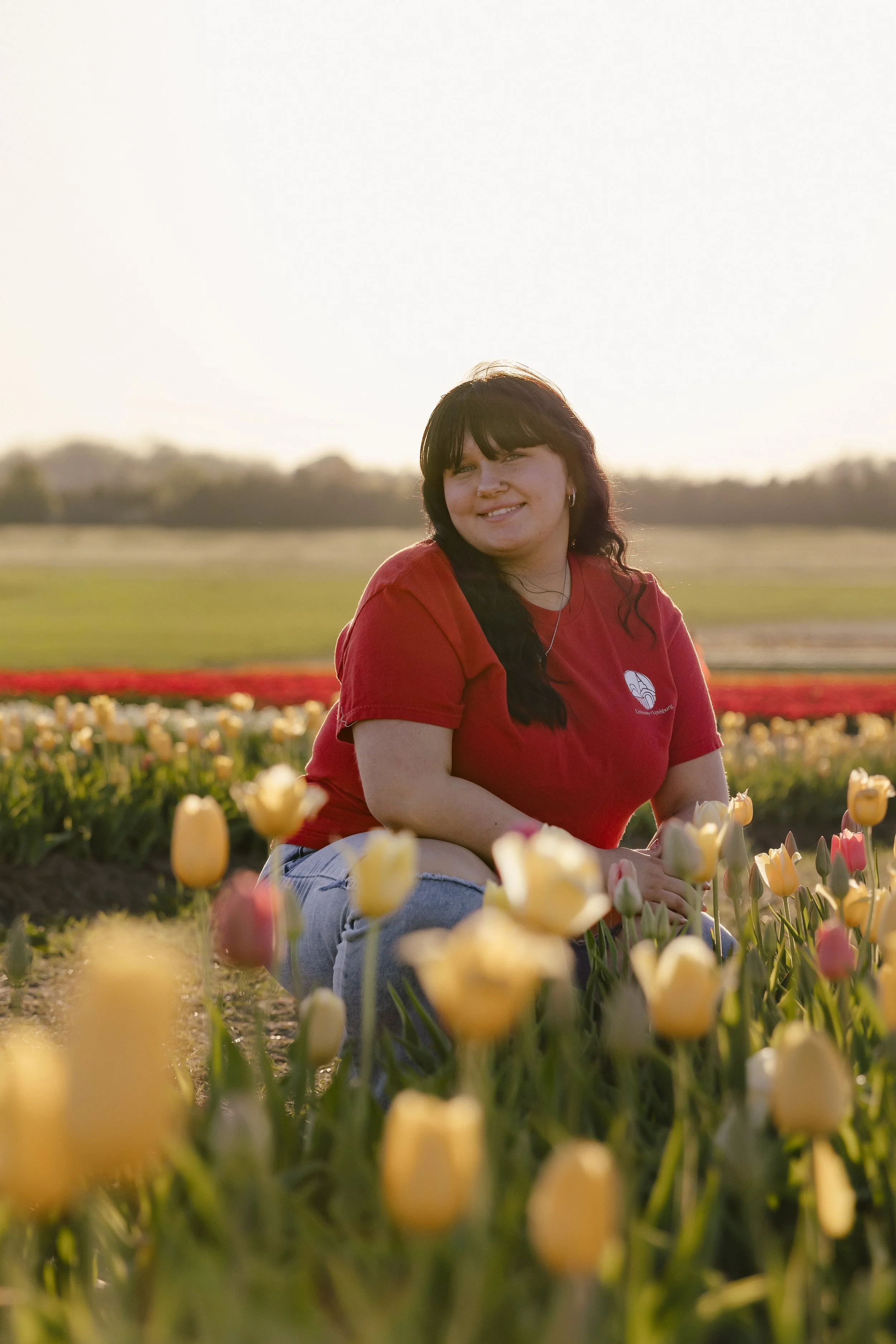 A young woman with dark hair and bangs, wearing a red t-shirt, sitting among blooming yellow and pink tulips in a field, with a soft blurred background of a wide field and a light sky during sunset or sunrise.