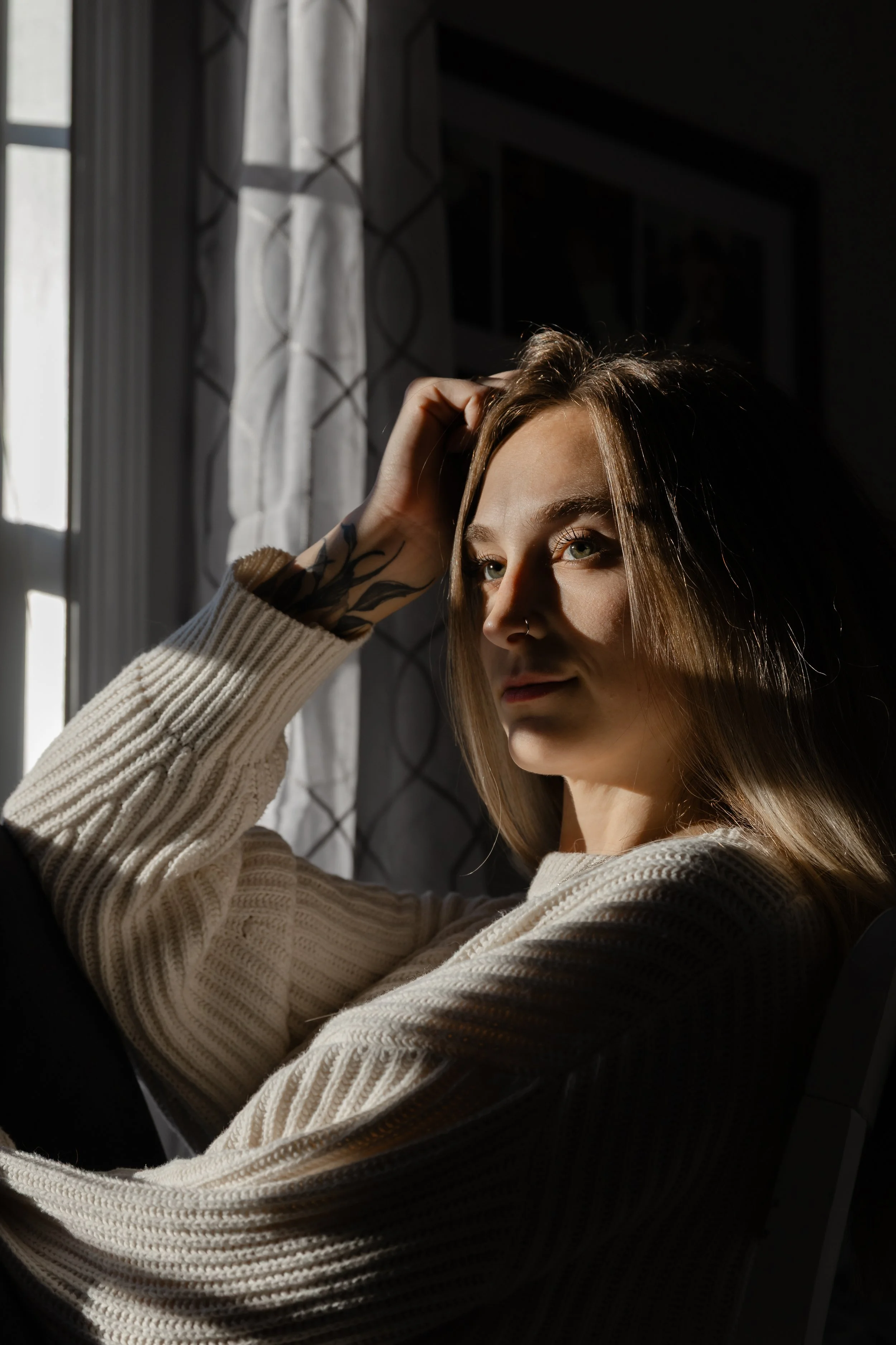 A young woman with light skin and long light brown hair, wearing a beige knit sweater, sitting near a window with sheer curtains, looking thoughtful as she catches sunlight on her face. She has a nose piercing and a tattoo on her left arm.