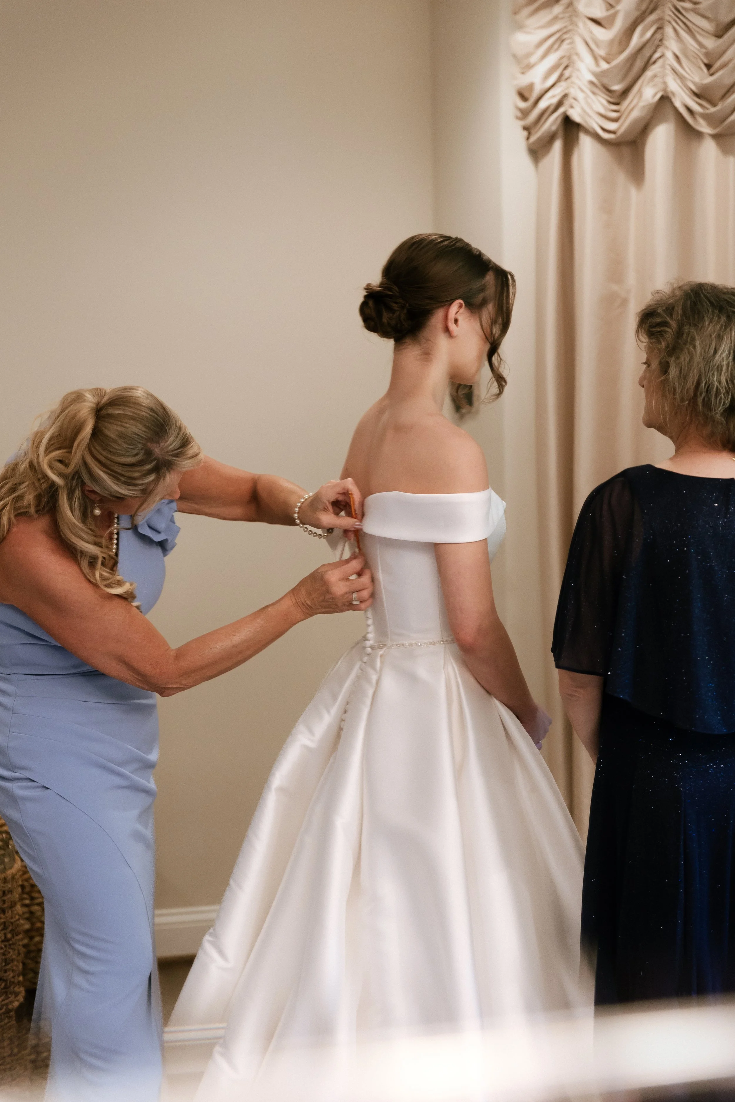 A bride in a white satin wedding gown is having her dress adjusted by an older woman, while another woman in a black dress stands nearby.