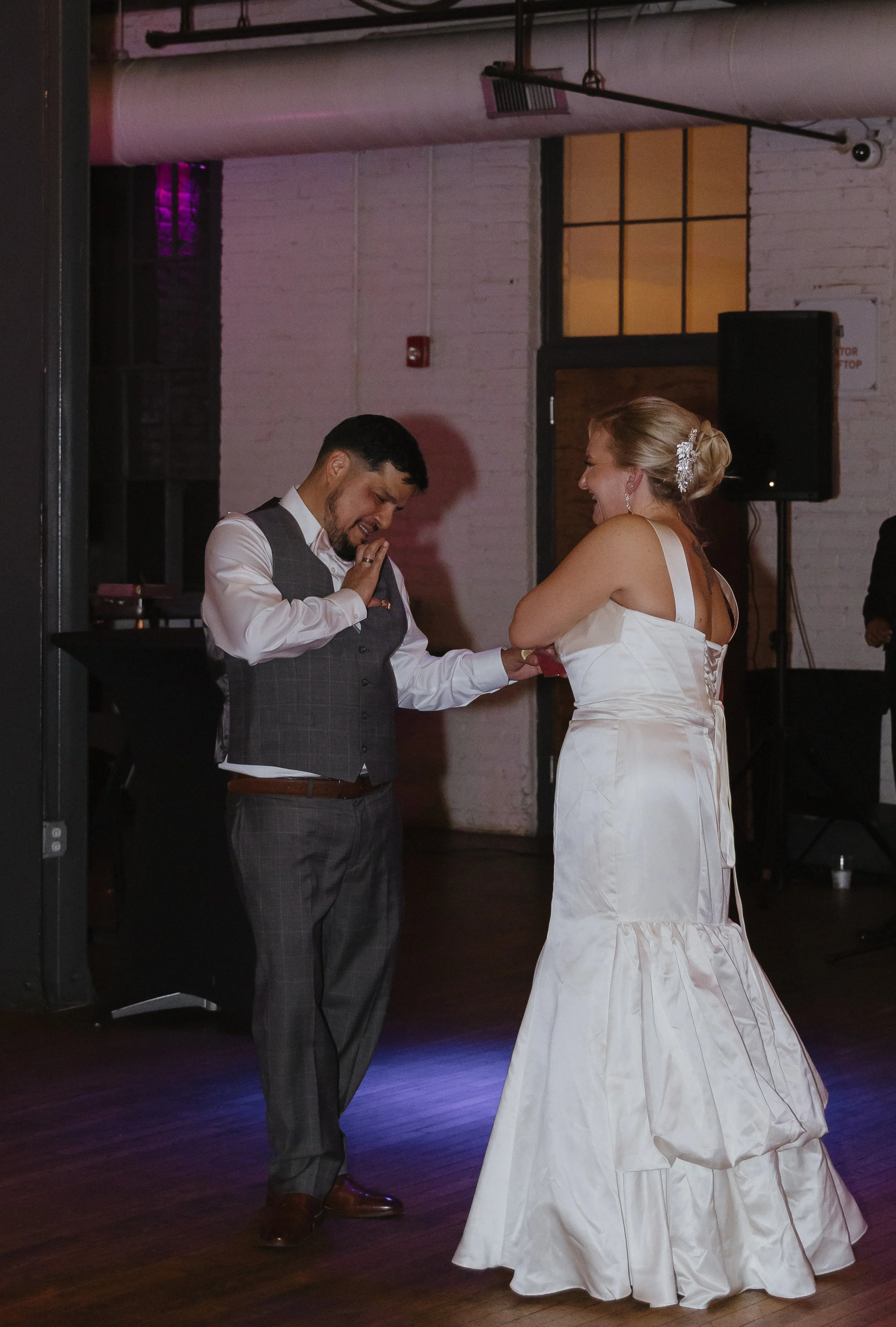 A bride and groom during their wedding dance, smiling and holding hands in a dimly lit venue with a DJ speaker in the background.