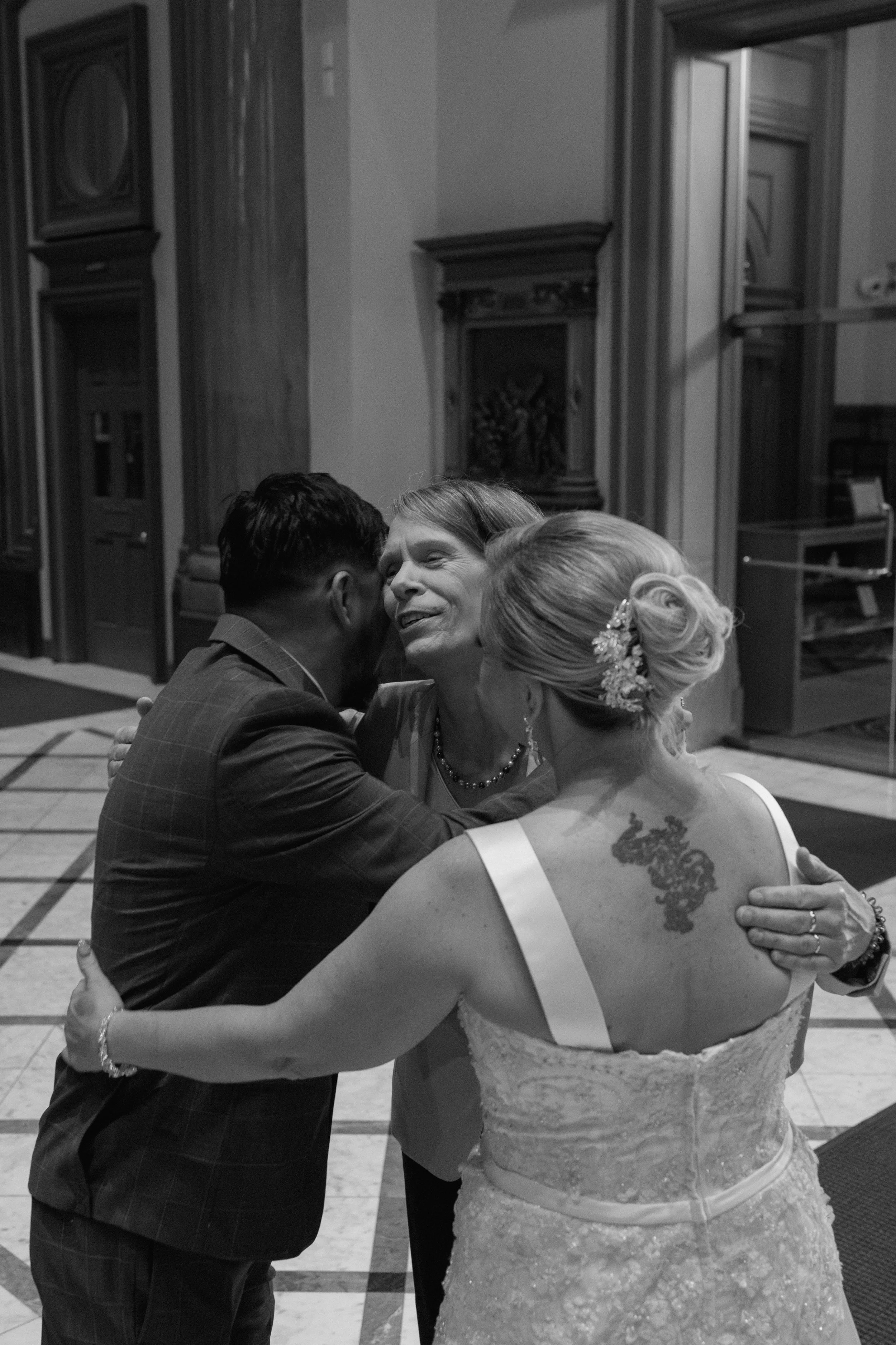 Three women hugging at a formal event in a grand hall, with one woman in a bride's gown, another woman wearing a light-colored blazer, and a man with a beard and short dark hair, all smiling and embracing.