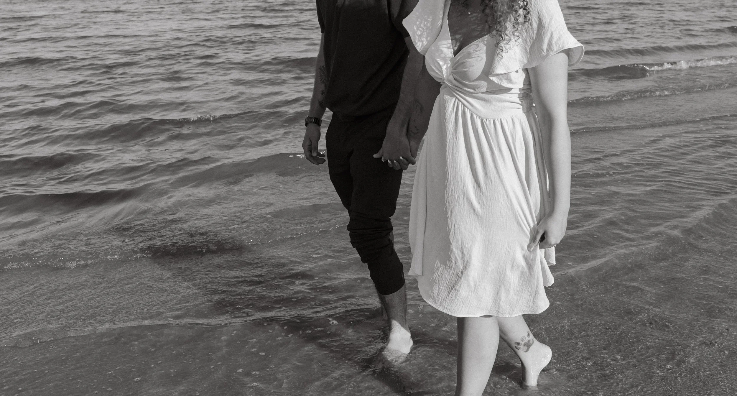 A black and white photo of a couple holding hands walking in the water at the beach. The man's face is not visible, wearing dark clothes. The woman wears a white dress, has curly hair, and has a tattoo on her ankle.