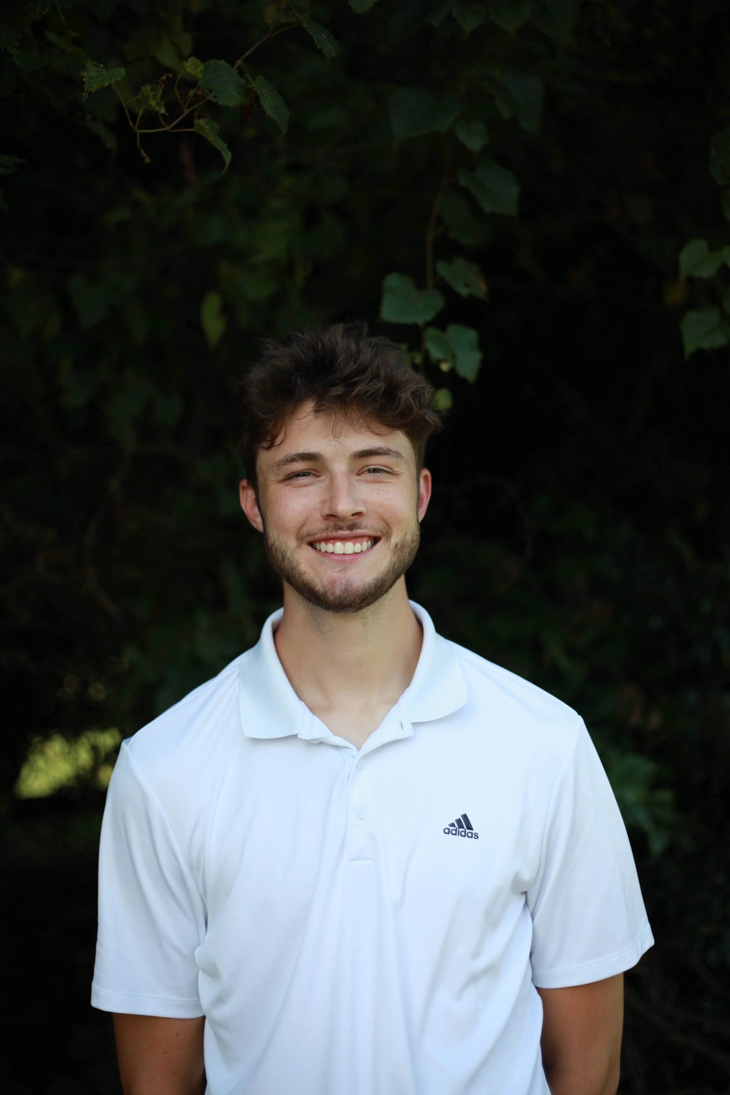 A young man smiling outdoors, wearing a white Adidas polo shirt, with a background of green foliage.