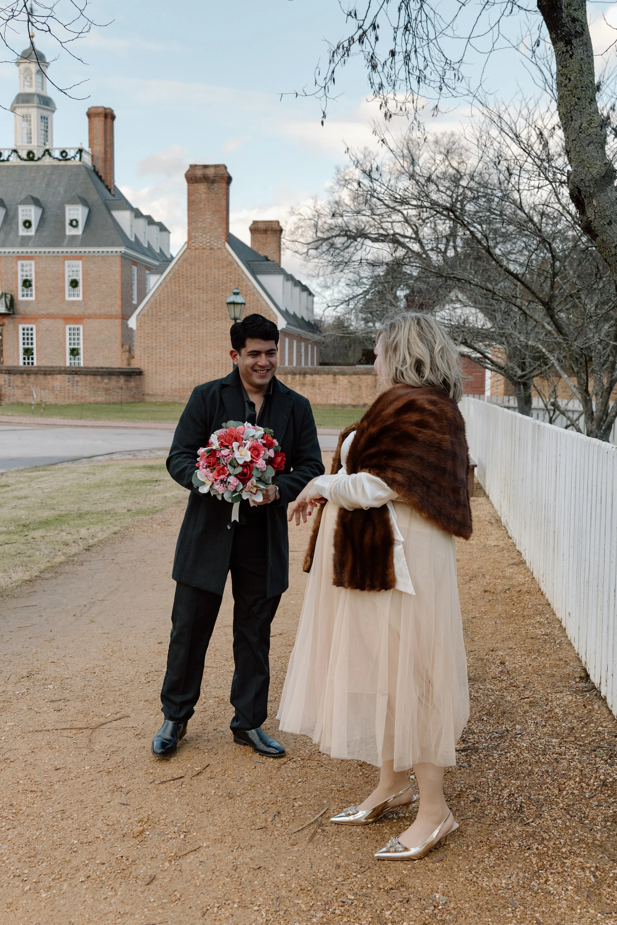 A man in a black coat holding a bouquet of pink, red, and white flowers is smiling and talking to a woman wearing a cream dress with a brown fur stole and gold shoes on an outdoor dirt path with a white picket fence and old brick buildings in the bac