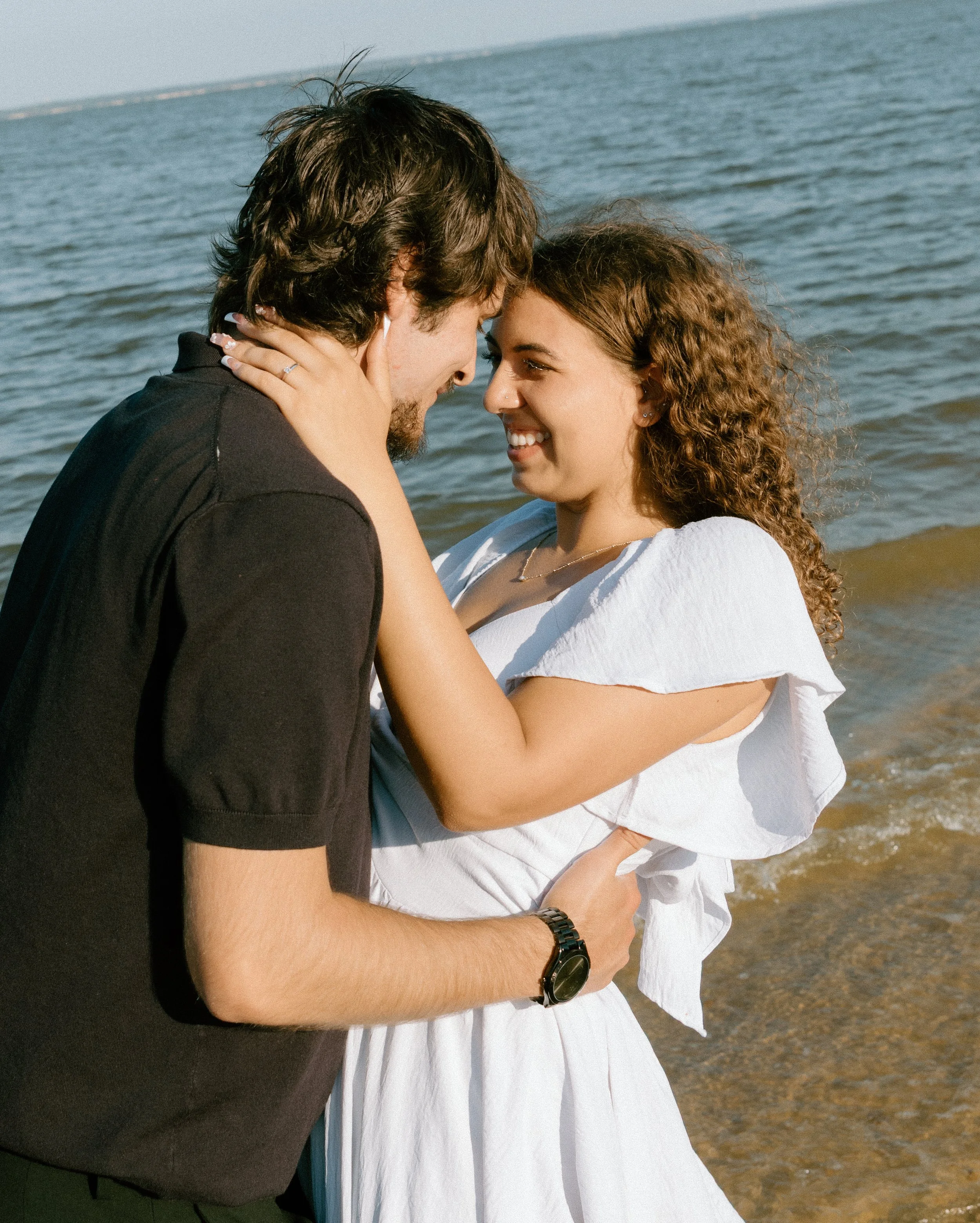 A couple is standing on a beach, holding each other and smiling, with the ocean and shoreline in the background.