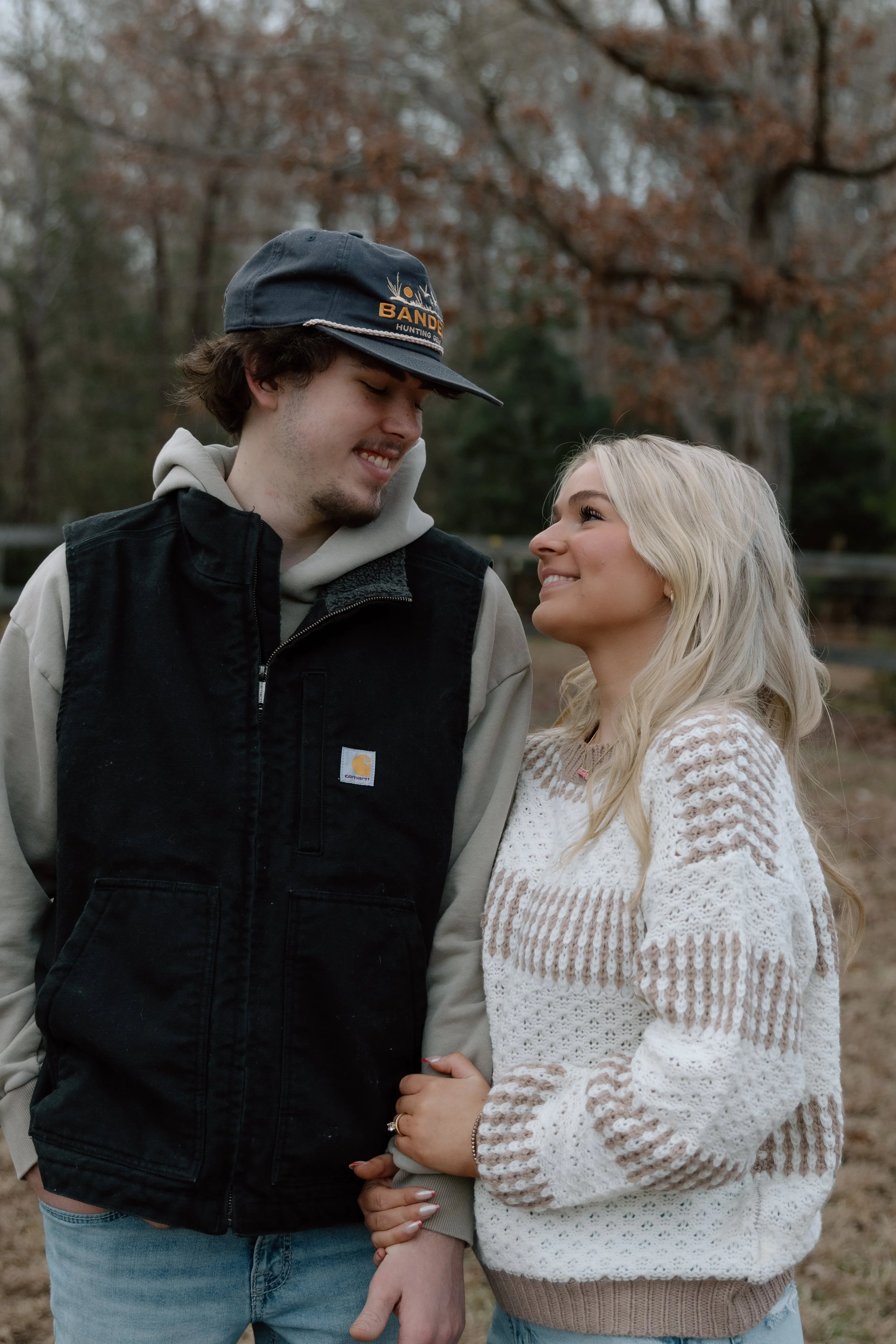 A young couple smiling and gazing at each other outdoors in autumn, holding hands with trees and a wooden fence in the background.