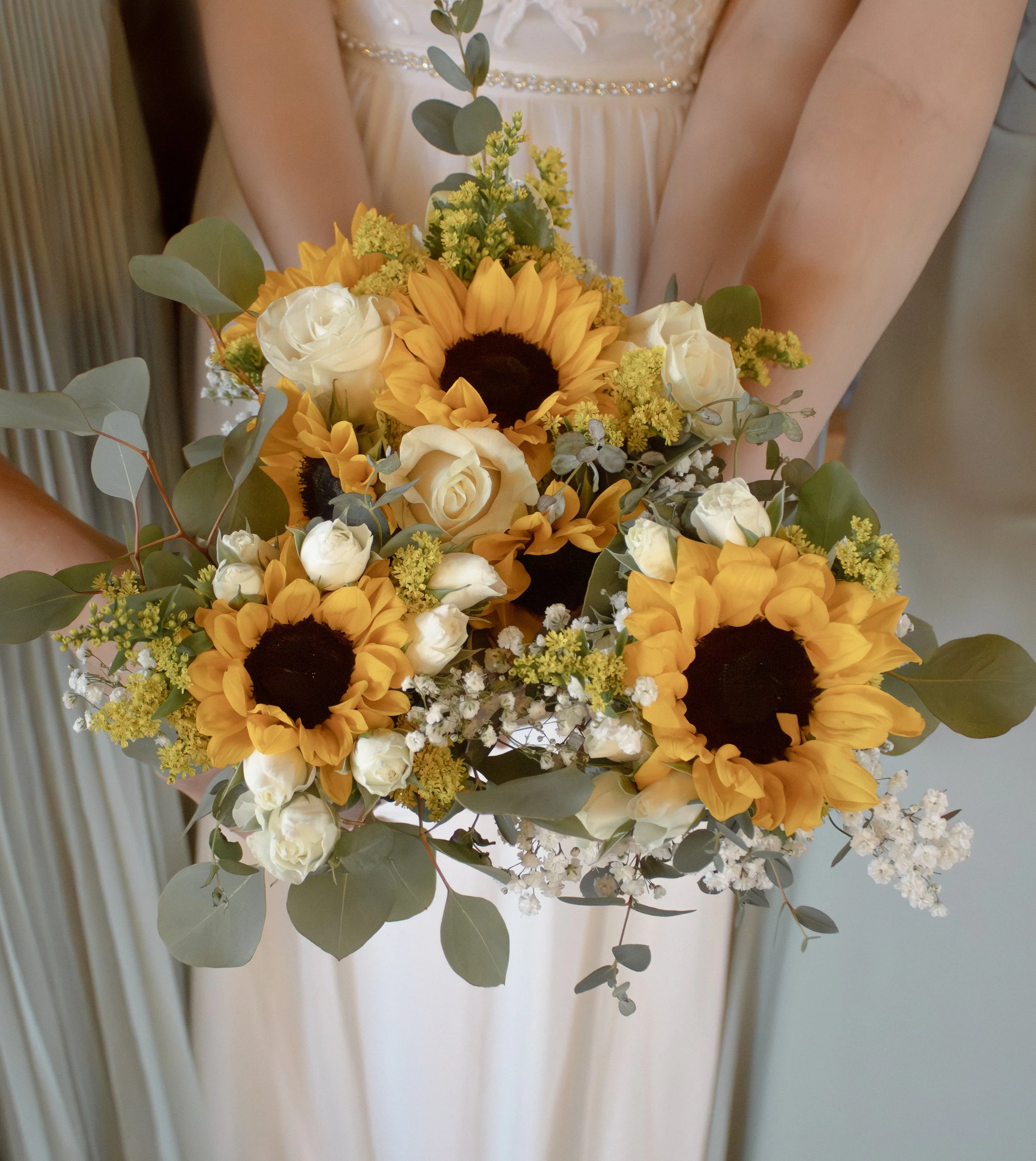 A floral bouquet consisting of yellow sunflowers, white roses, small yellow flowers, and green eucalyptus leaves, held against a cream-colored dress with embroidery and bead details.